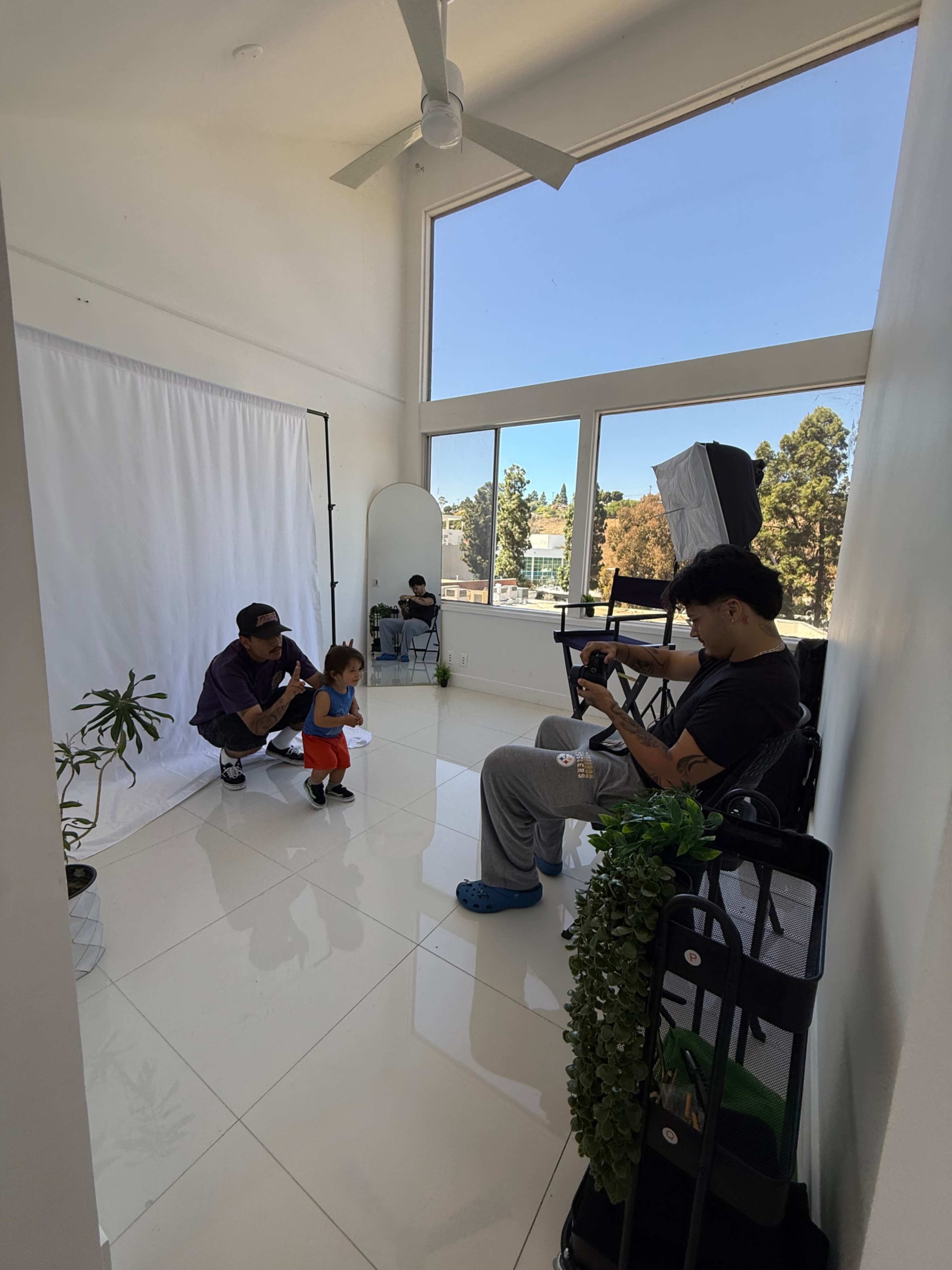 A child interacts with an adult in a well-lit studio while another adult sits nearby, observing and using a phone.