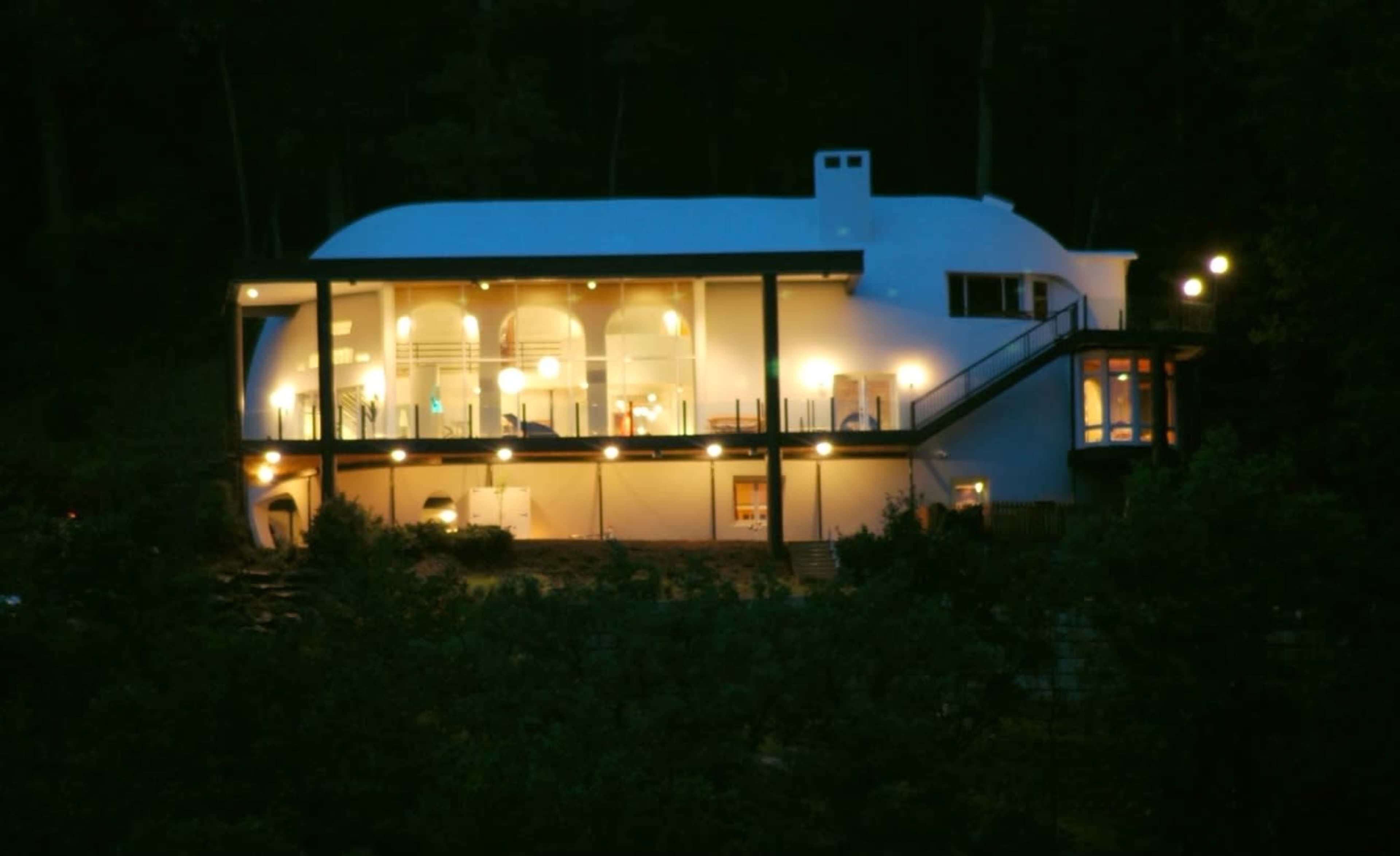 A modern, white, rounded house is illuminated at night, featuring multiple balconies and large windows overlooking a wooded area.