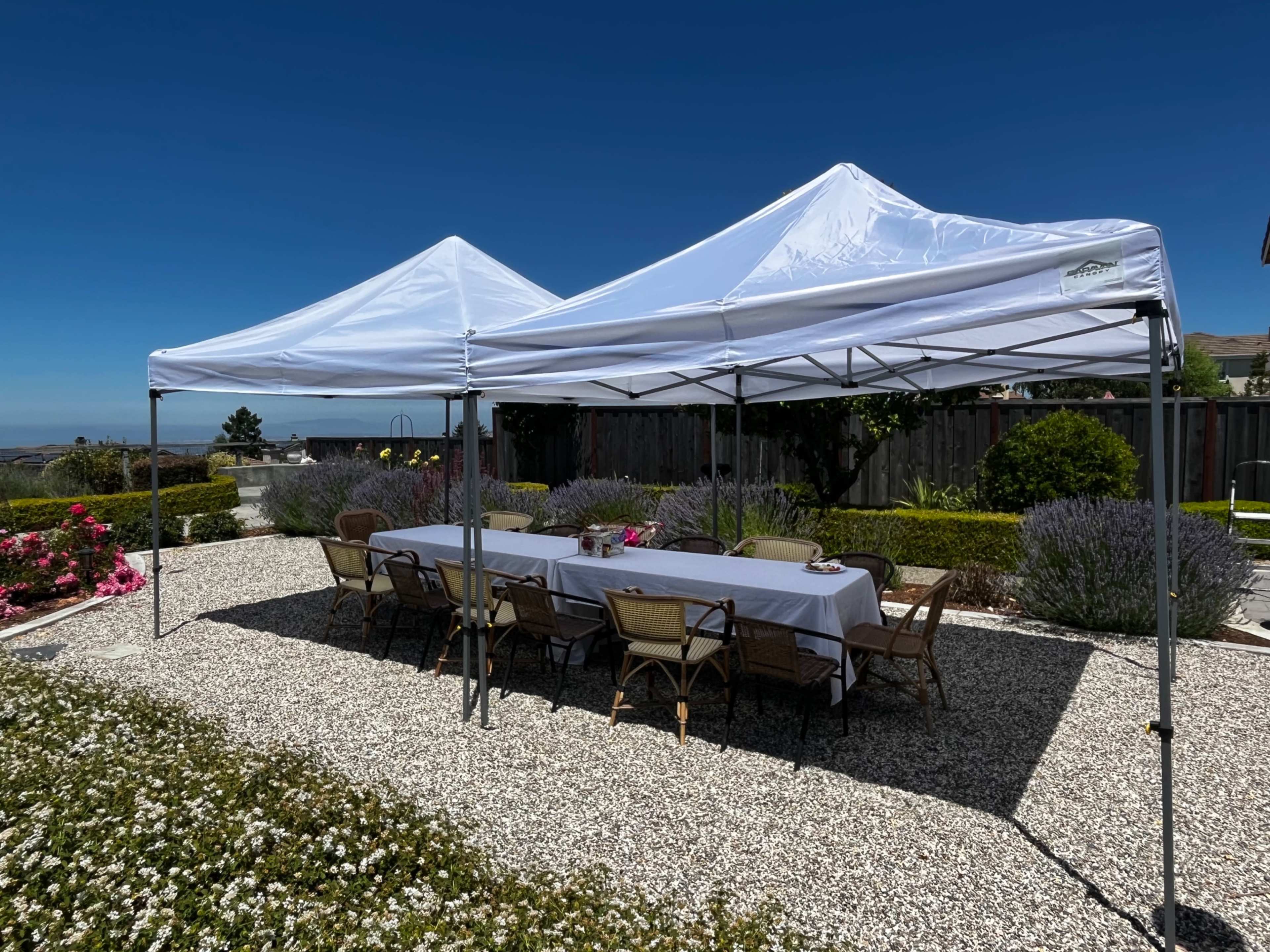 Two white canopies are set up over a long table surrounded by chairs on a gravel patio with landscaping in the background.