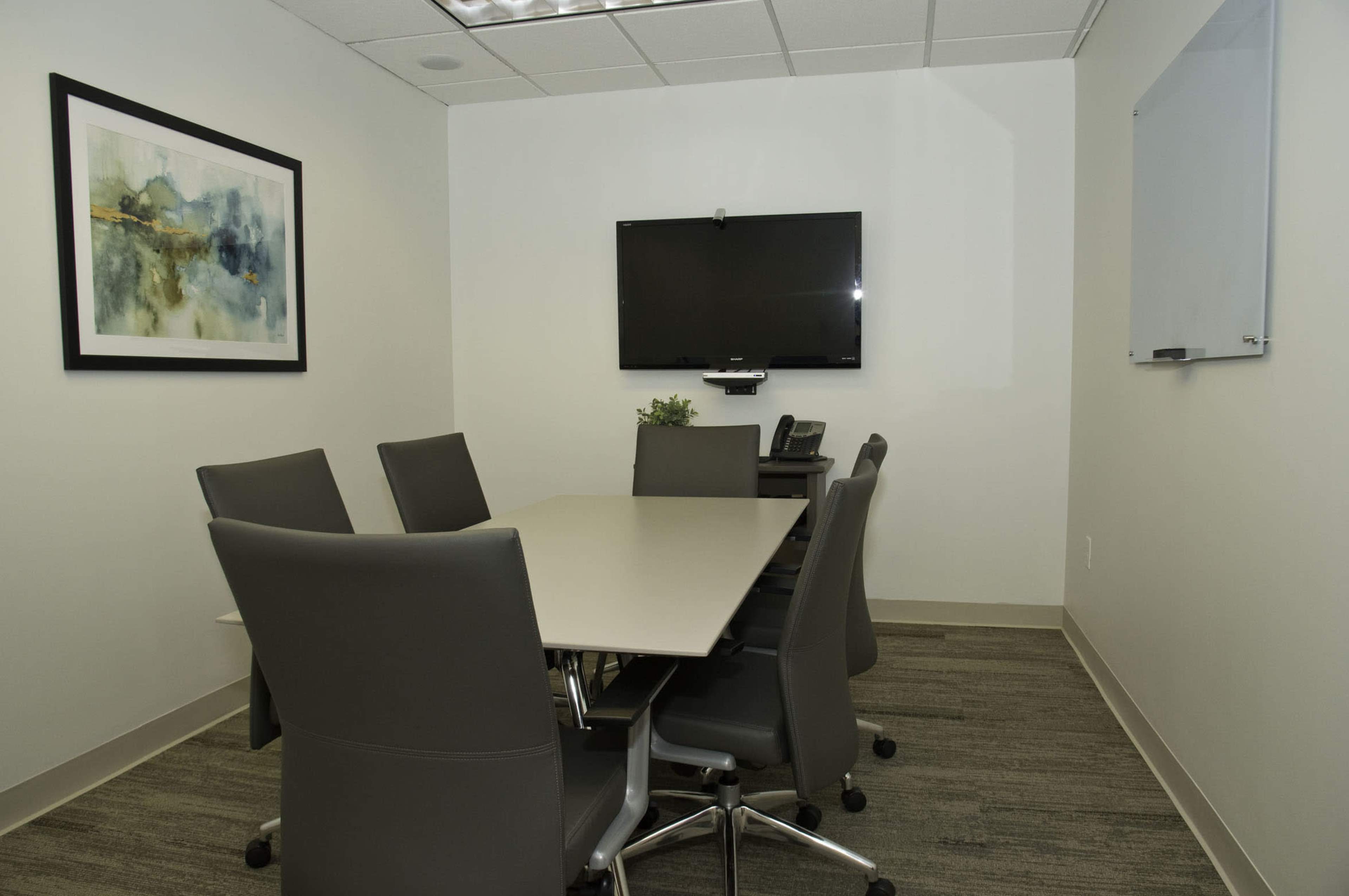 The image features a small conference room with a rectangular table surrounded by six chairs, a wall-mounted television, and a framed artwork.