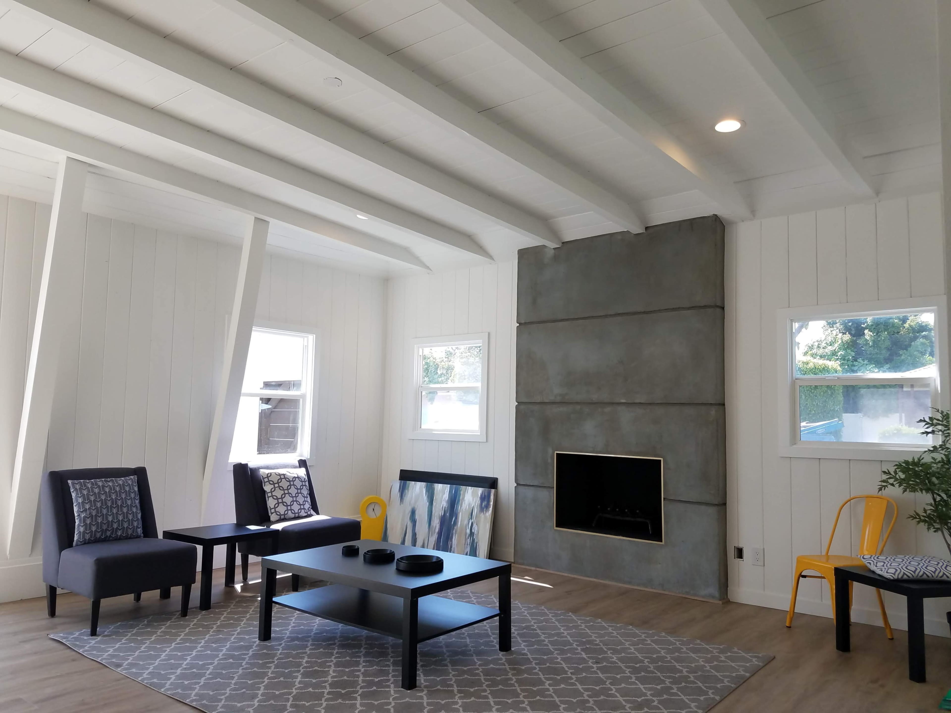 A modern living room with a concrete-featured fireplace, a dark coffee table, and two armchairs, all set against a backdrop of white walls and exposed beams.