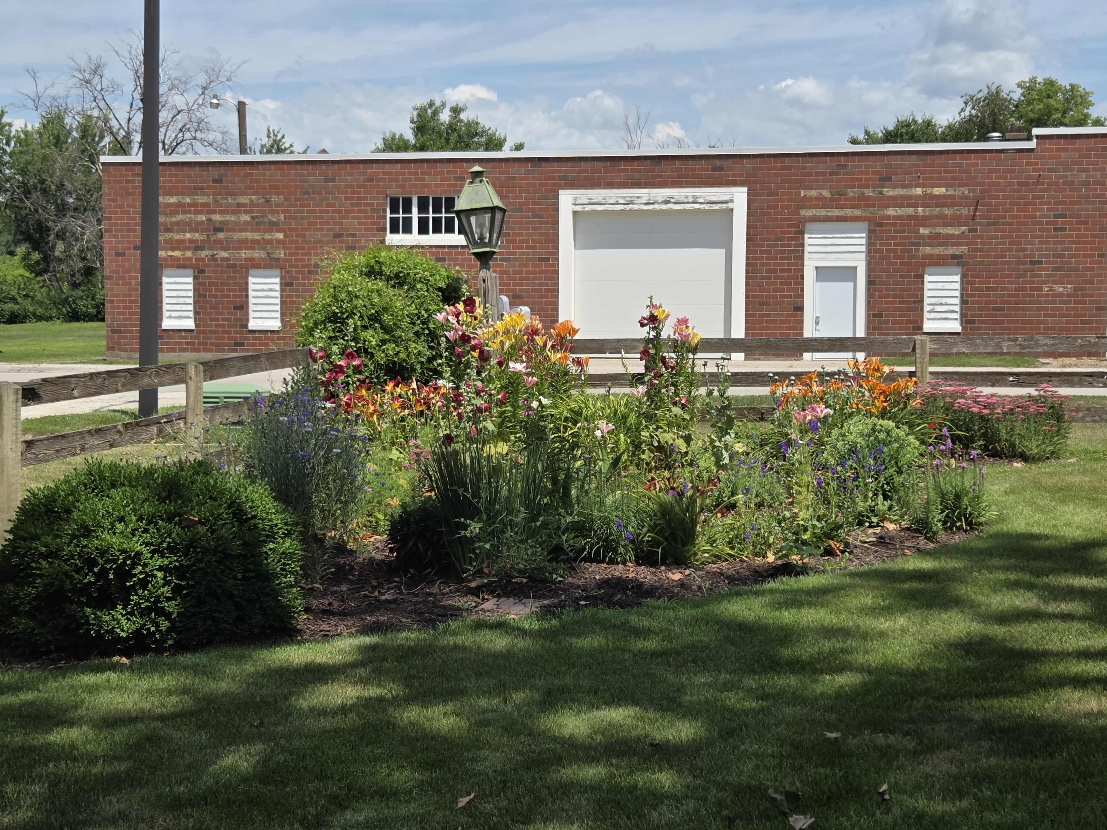 A flowerbed filled with various blooming plants is located in front of a red brick building with a garage door.