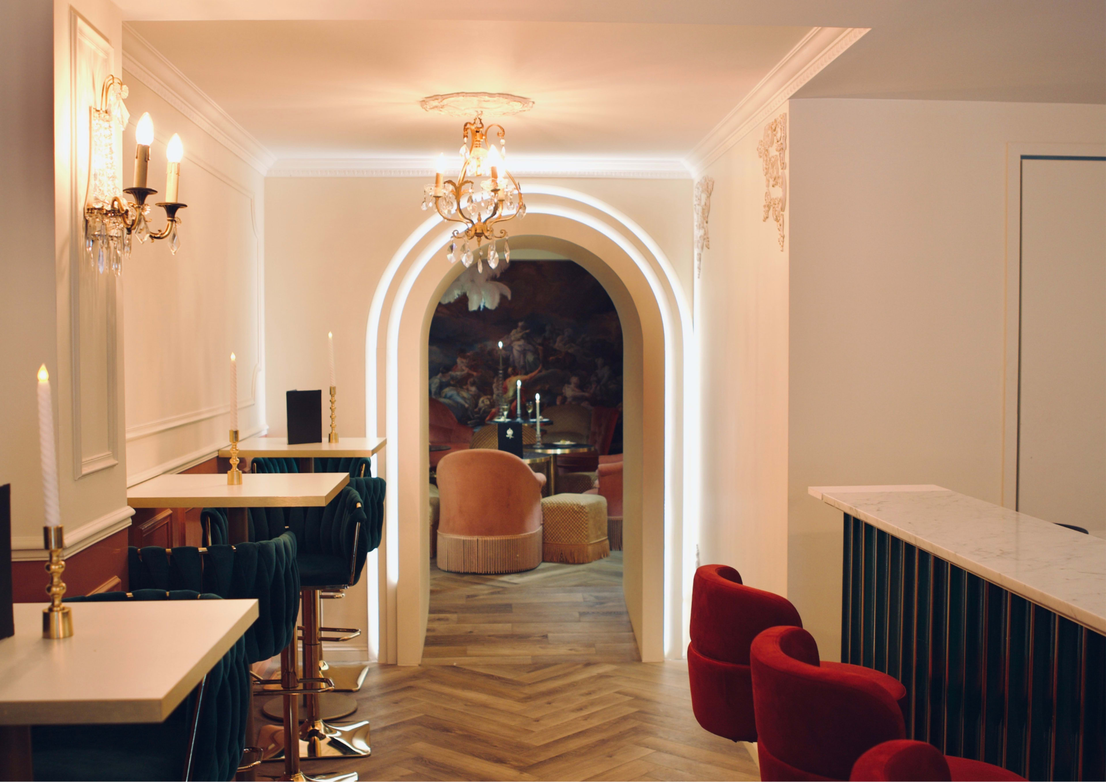 The image shows a well-lit hallway with arches leading to a cozy seating area, flanked by modern bar stools and tables on one side.