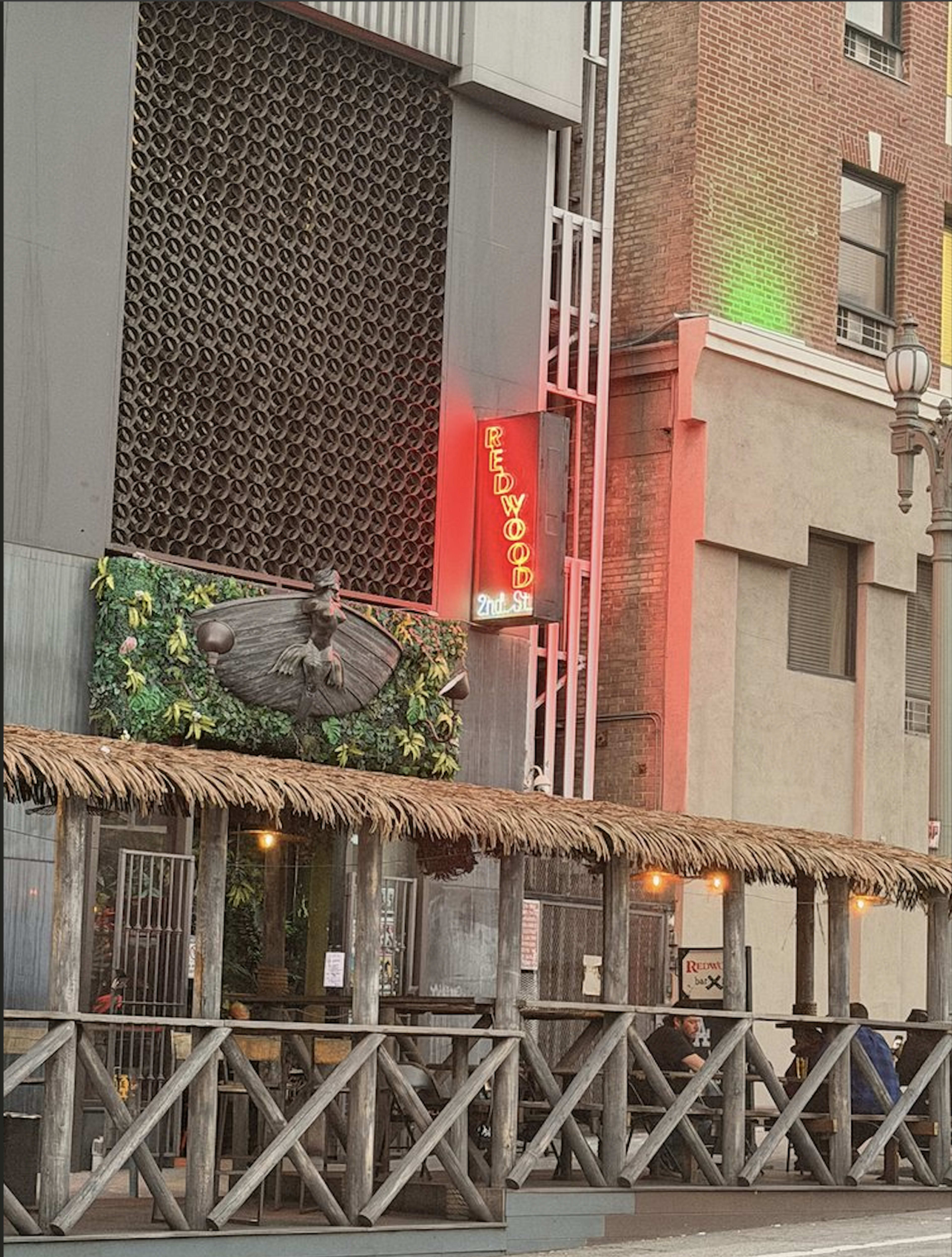 The image shows a bar or restaurant with a thatched-roof outdoor seating area, illuminated by neon lights that read "Redwoods," located in an urban setting.