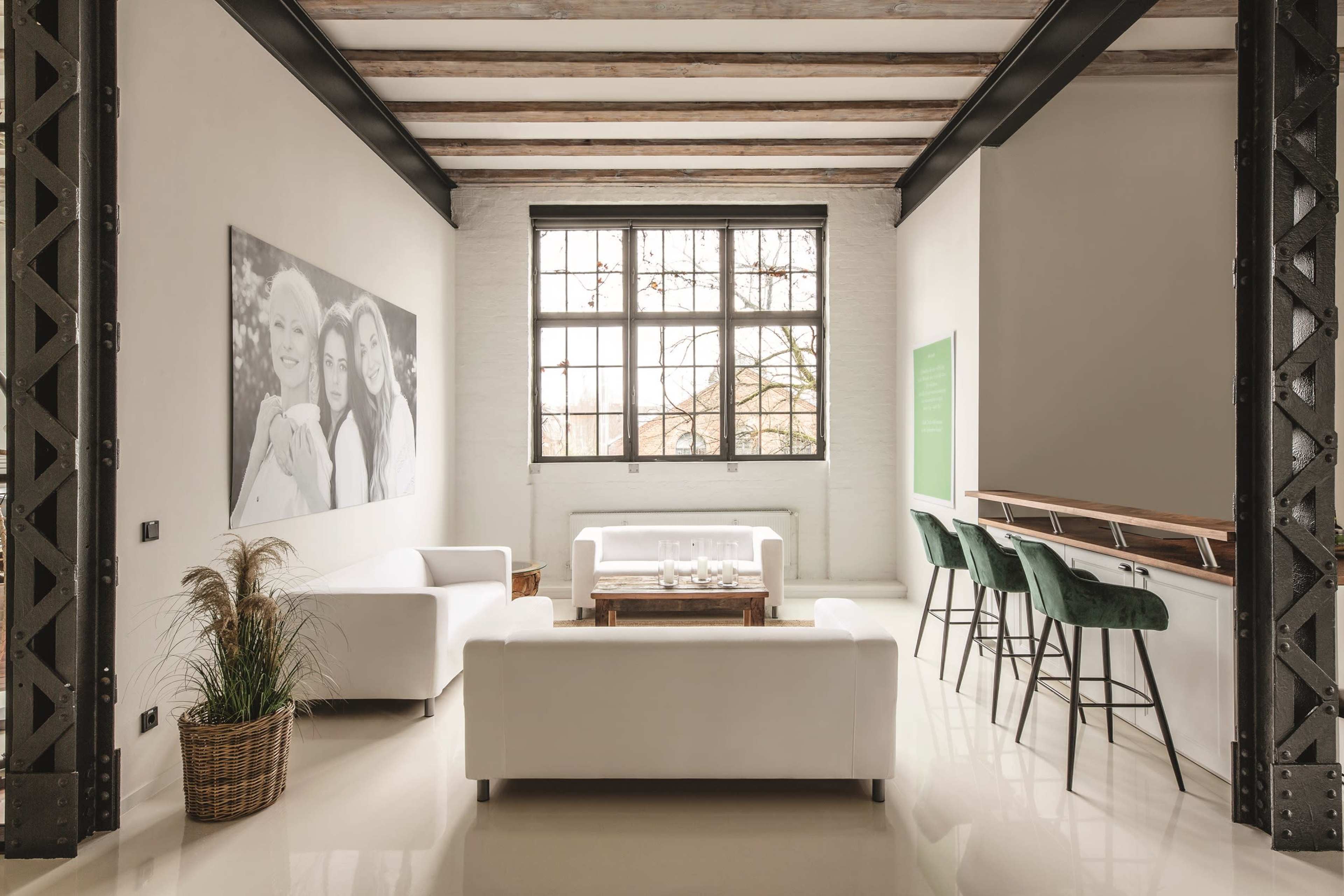 The image shows a minimalist living space with two white sofas, a wooden coffee table, bar seating with green stools, and large windows allowing natural light.