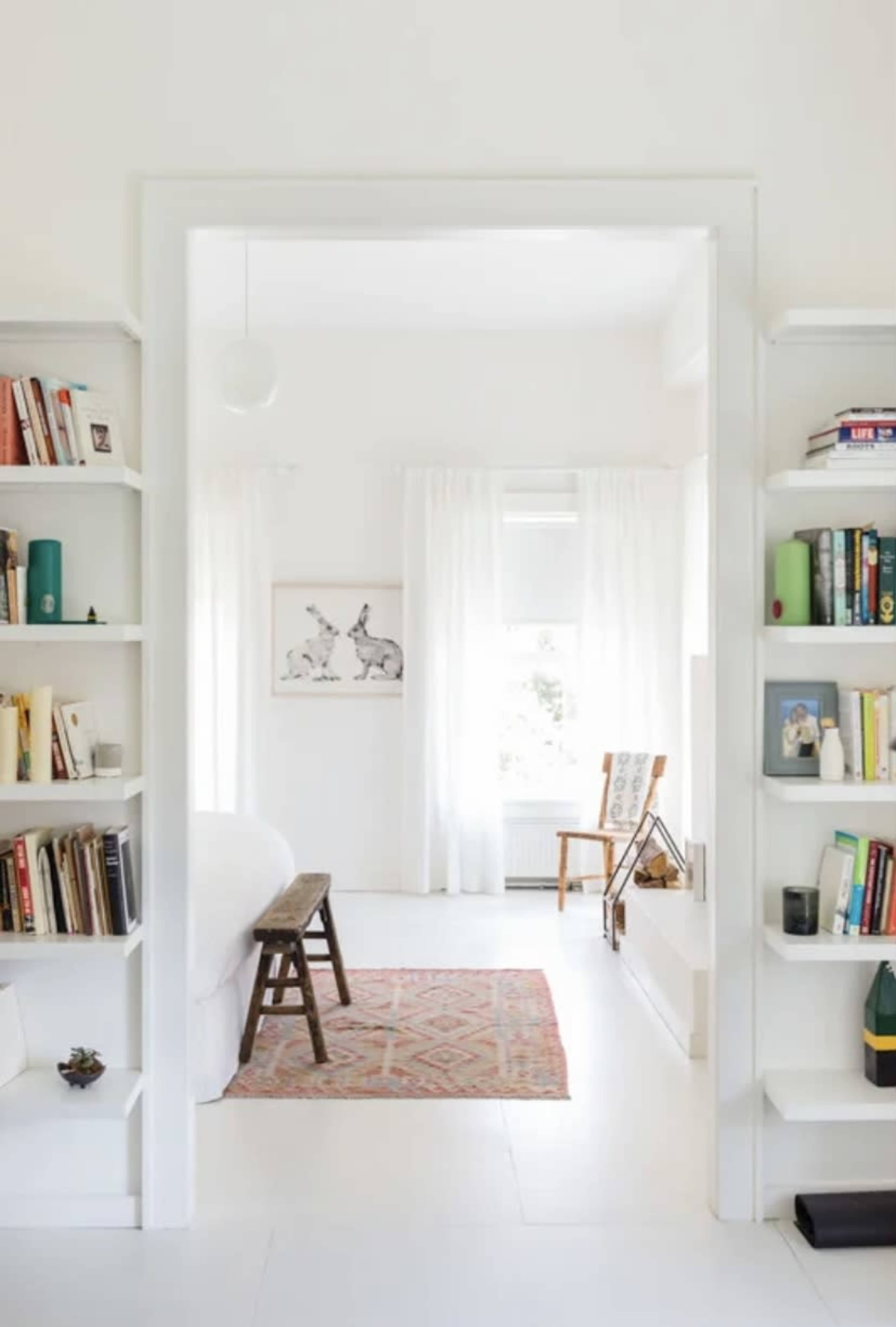 A bright, minimalist room features a doorway framed by bookshelves, leading to a space with a bed, a patterned rug, and a chair by a window.