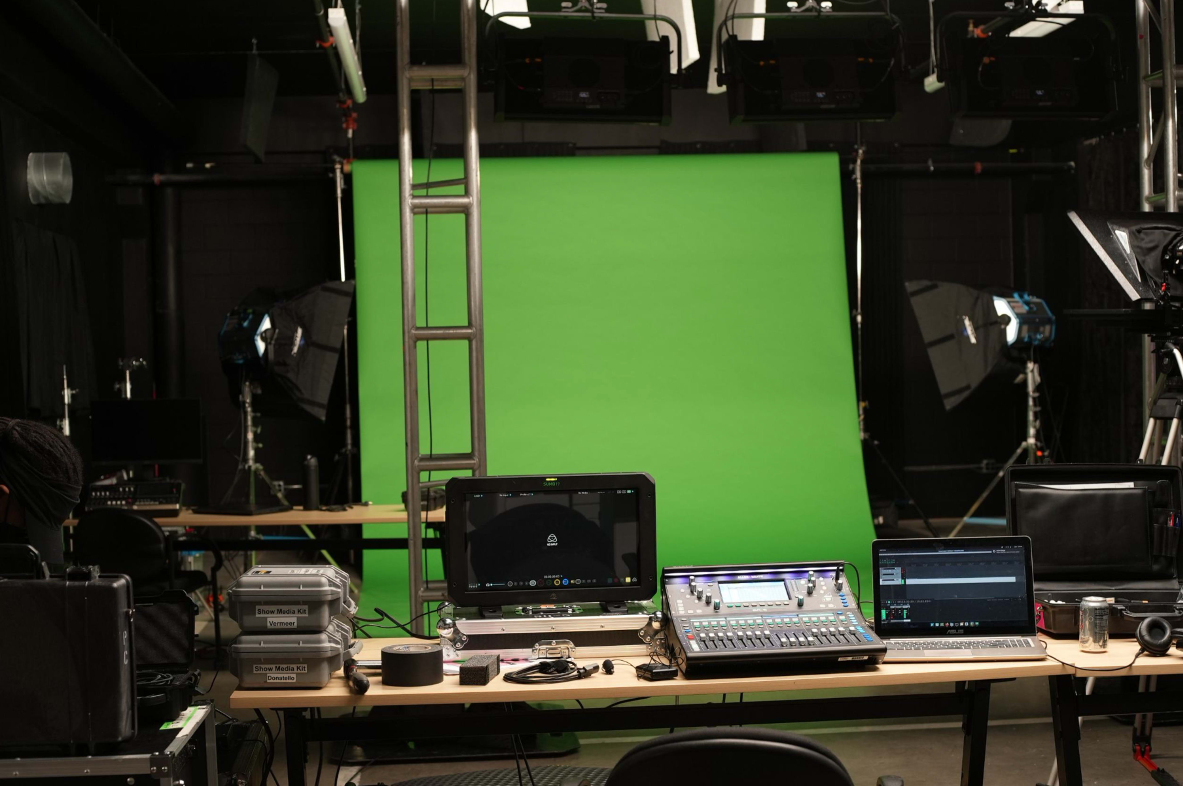 A production studio with a green screen setup, flanked by lighting equipment and recording devices on a worktable.