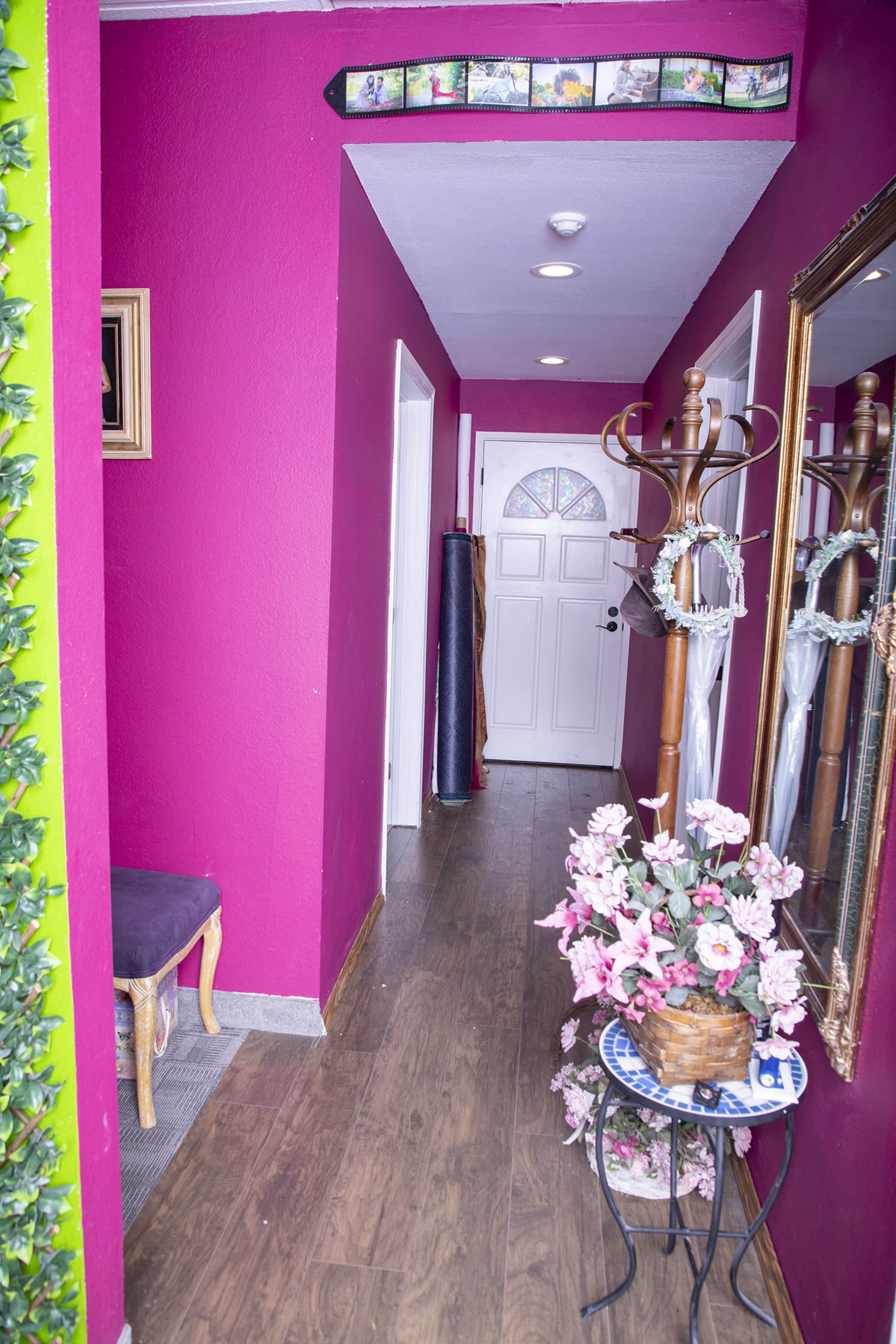A hallway with pink walls, a wooden floor, a coat rack, a mirror, a decorative plant, and a door at the far end.