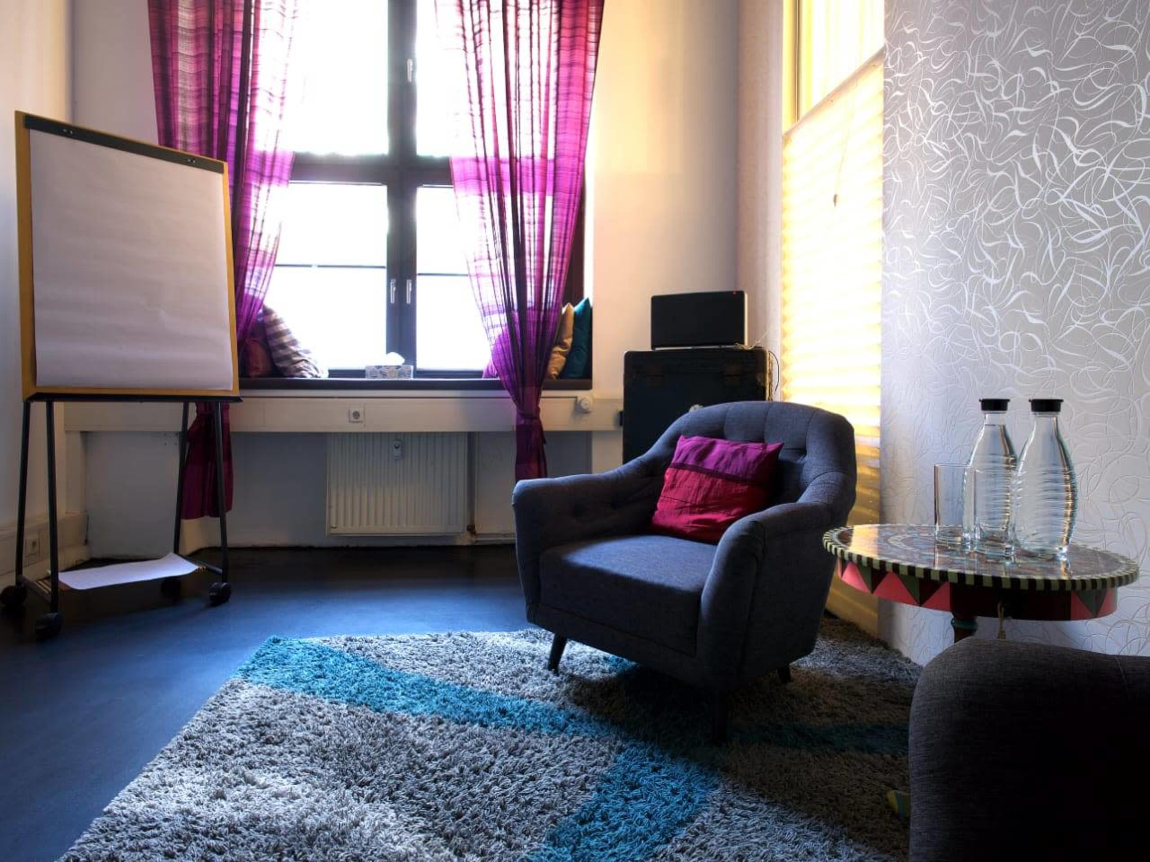 A cozy room features a gray armchair, a table with glass bottles, a flip chart stand, and vibrant curtains by a large window.