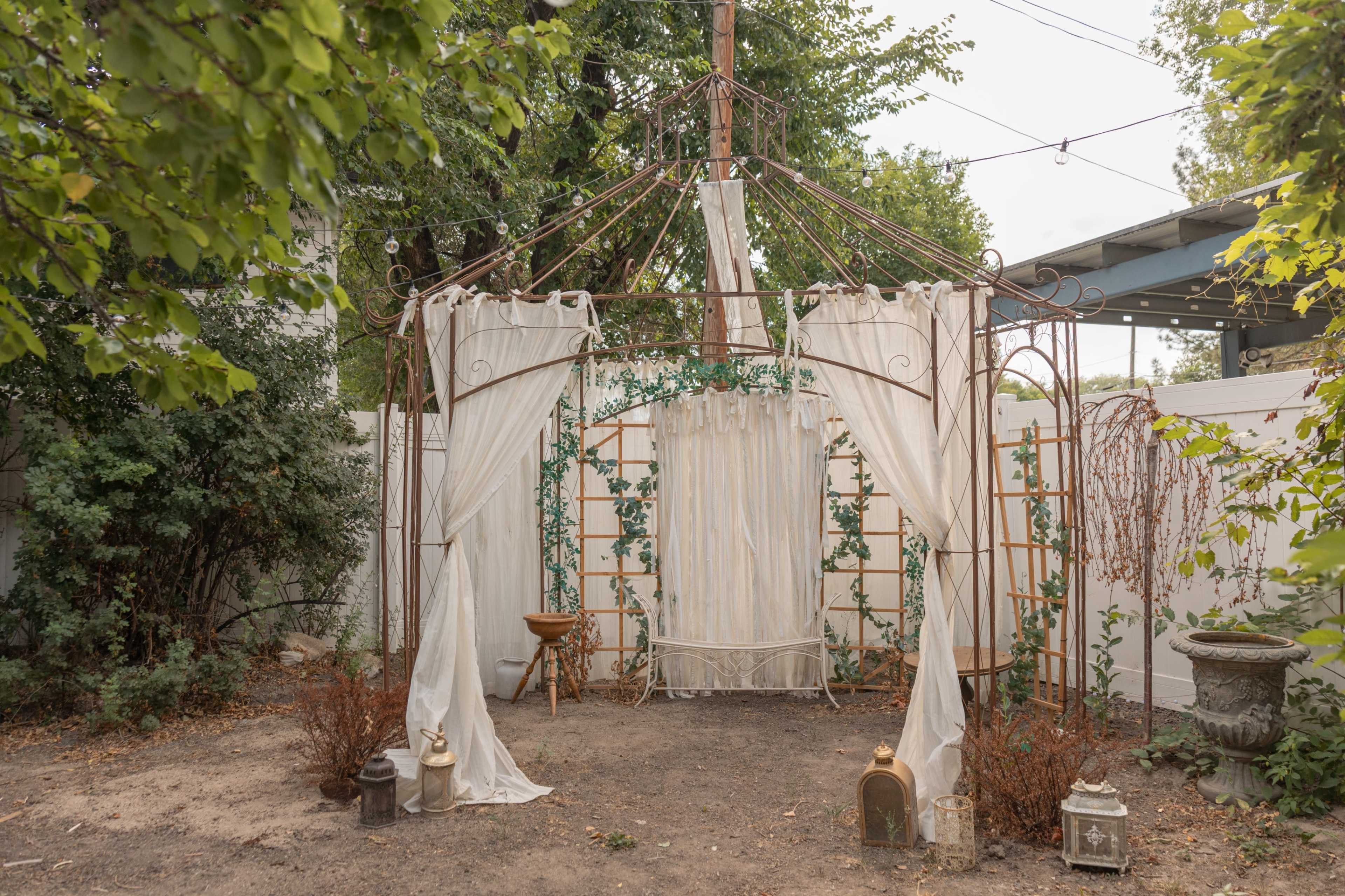 The image shows a decorative outdoor gazebo surrounded by greenery, with fabric draped elegantly and lanterns placed around the floor.