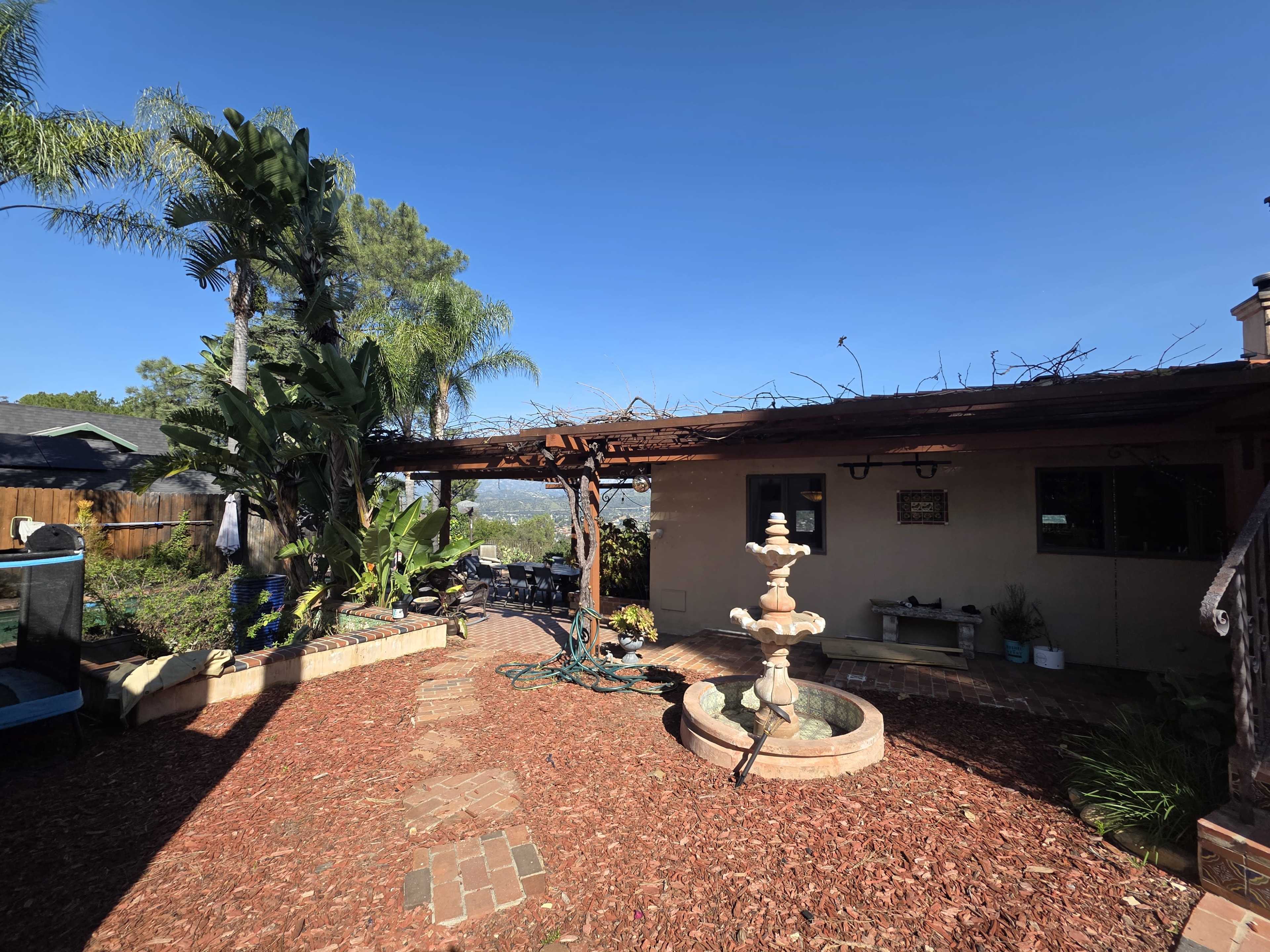 The image shows a backyard with a fountain at its center, surrounded by a pathway made of bricks and landscaping, under a clear blue sky.