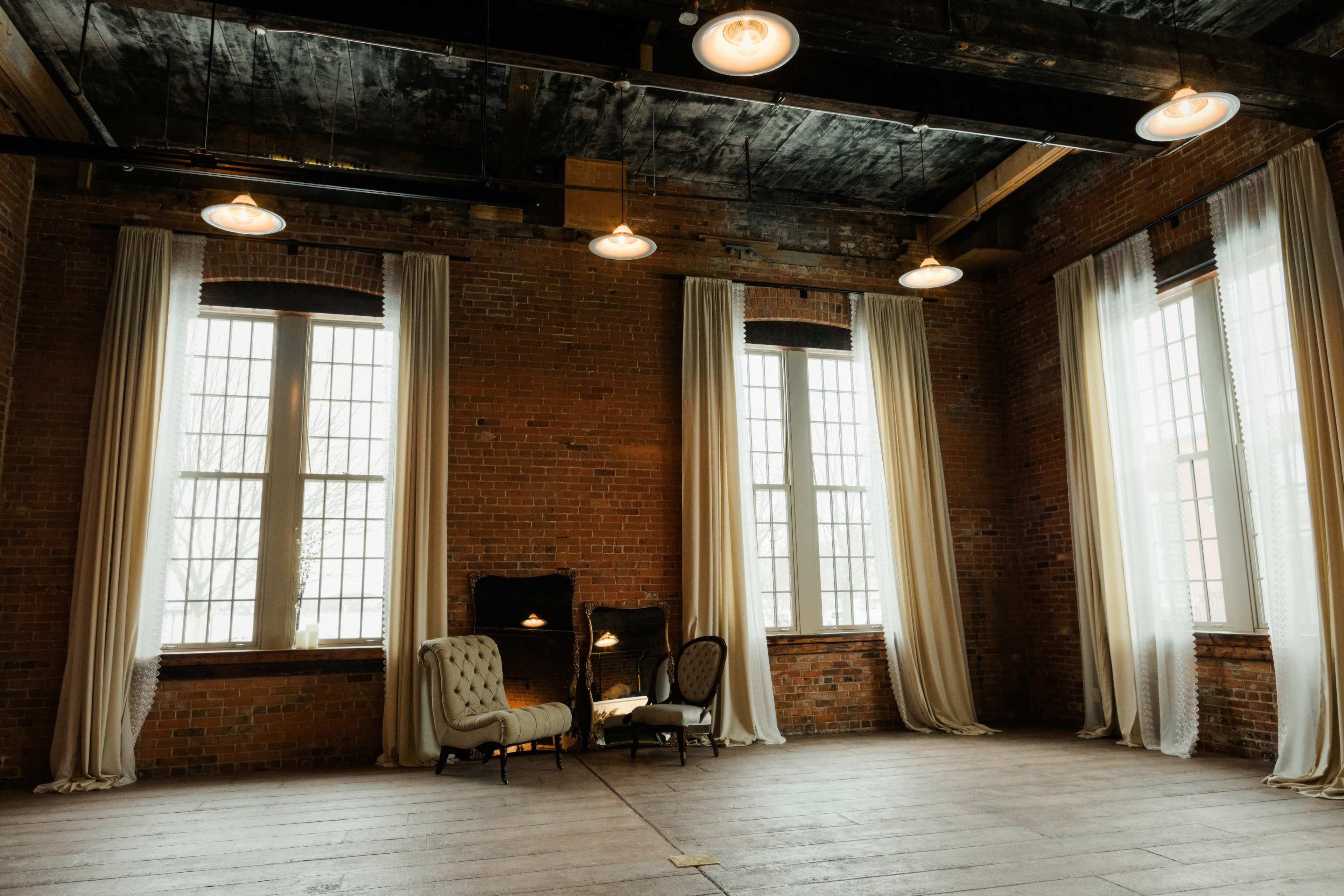 The spacious room features large windows, exposed brick walls, and a seating area with a vintage sofa and chairs.