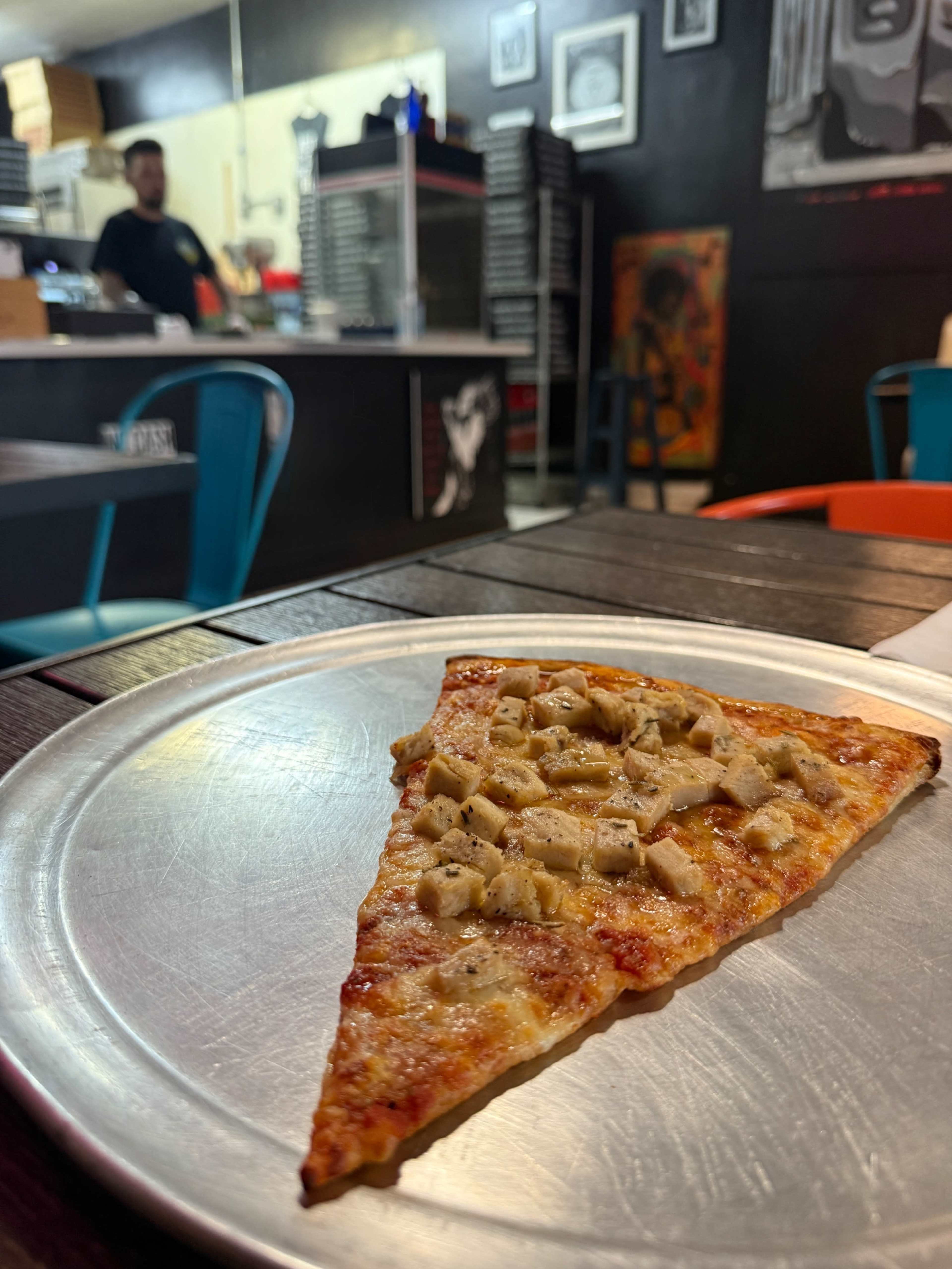 A slice of pizza topped with chicken sits on a metal plate in a pizza restaurant, with a blurred figure working in the background.