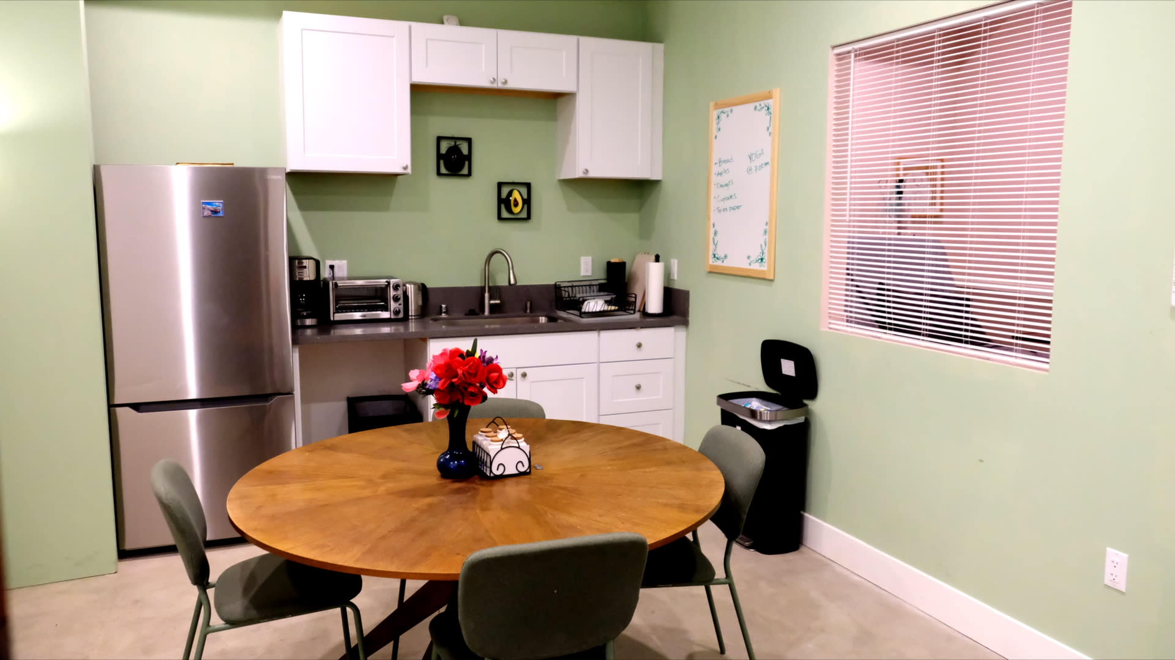 A kitchen with green walls features a round wooden table, a stainless steel refrigerator, and various kitchen appliances on a countertop.