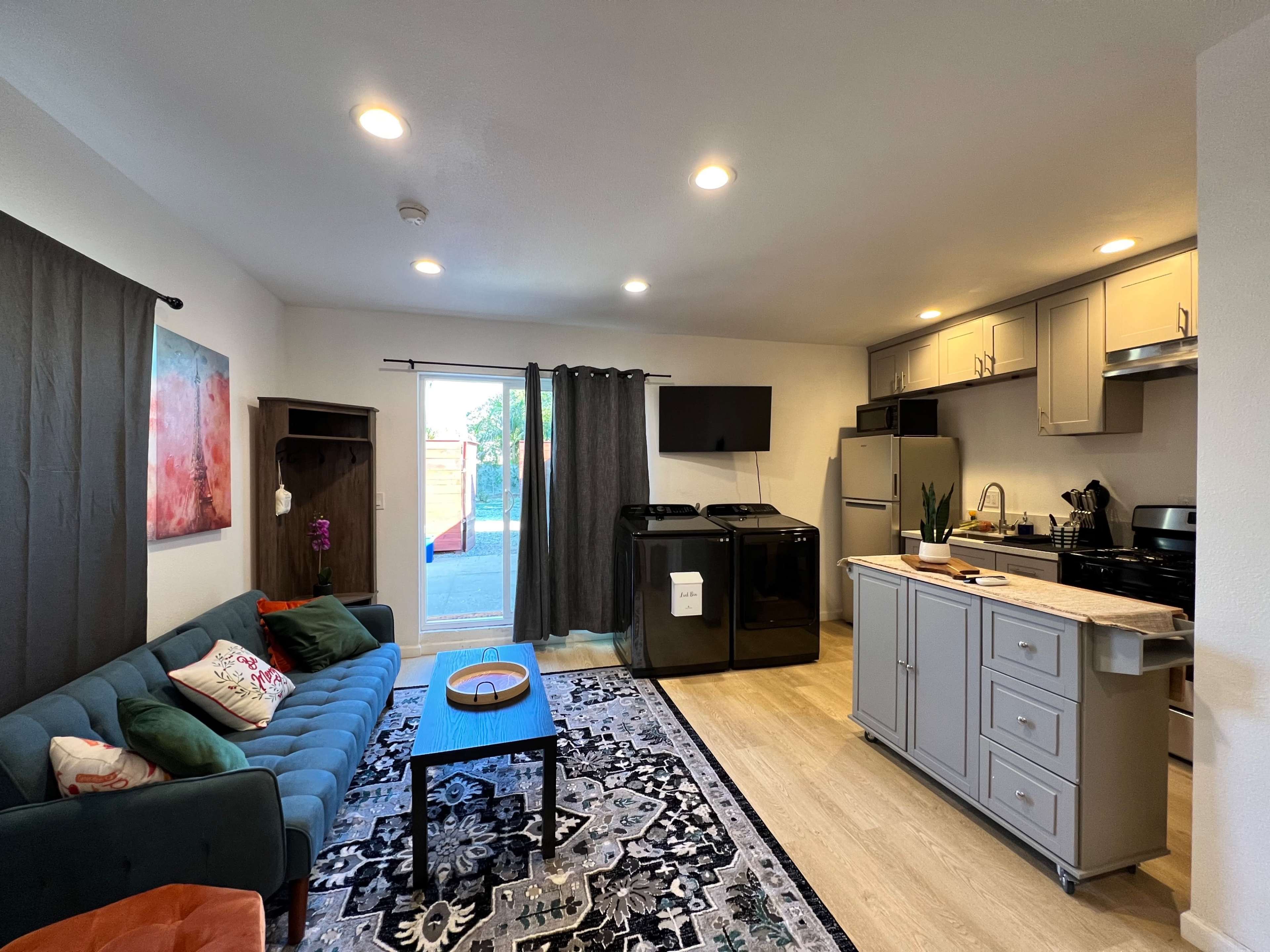 A modern open-concept living space featuring a kitchen with gray cabinets, a black refrigerator, and a comfortable seating area with a teal couch and decorative rug.
