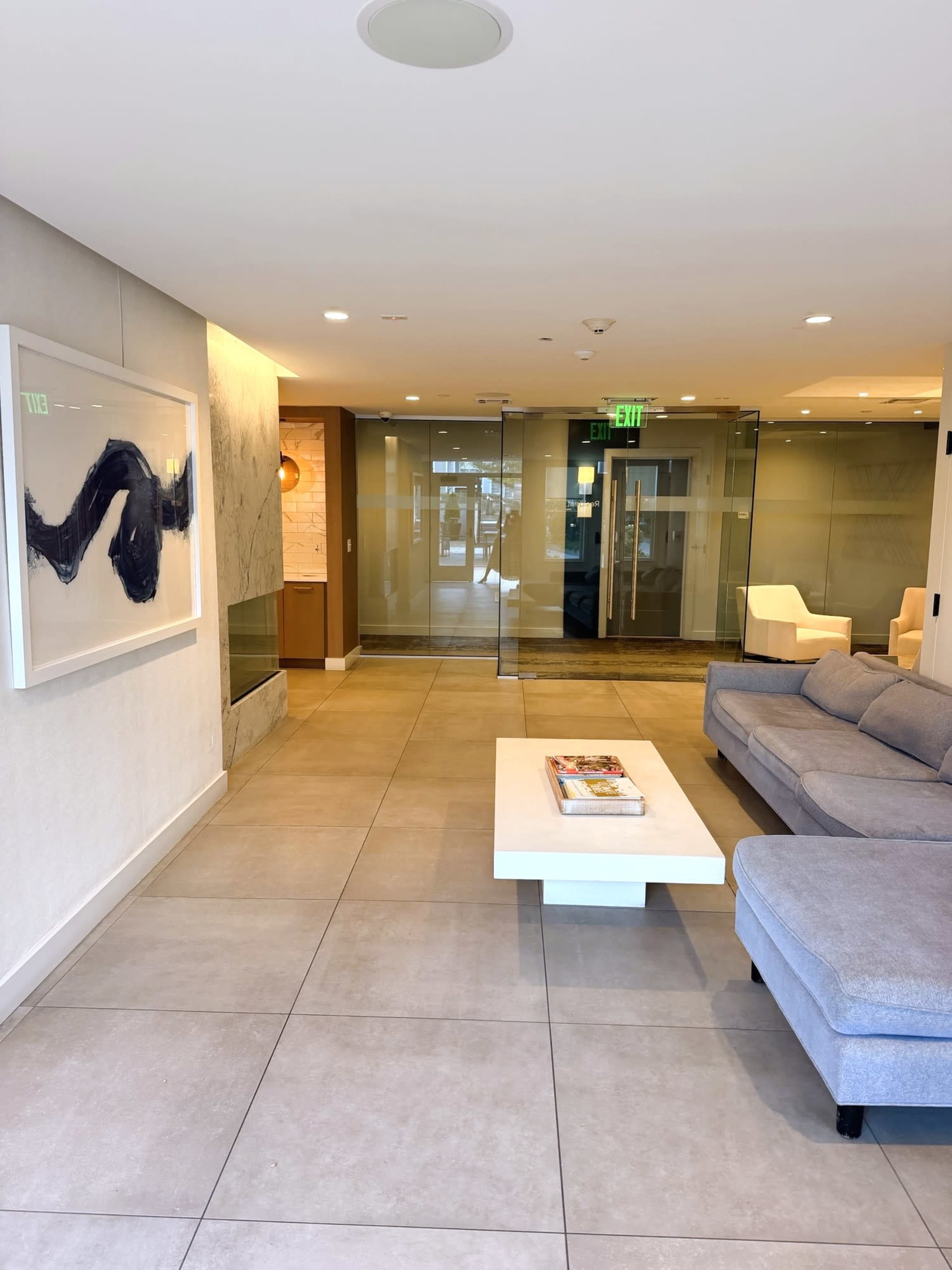 The image shows a modern lobby area featuring a gray couch, a white coffee table, and glass doors leading to another space.
