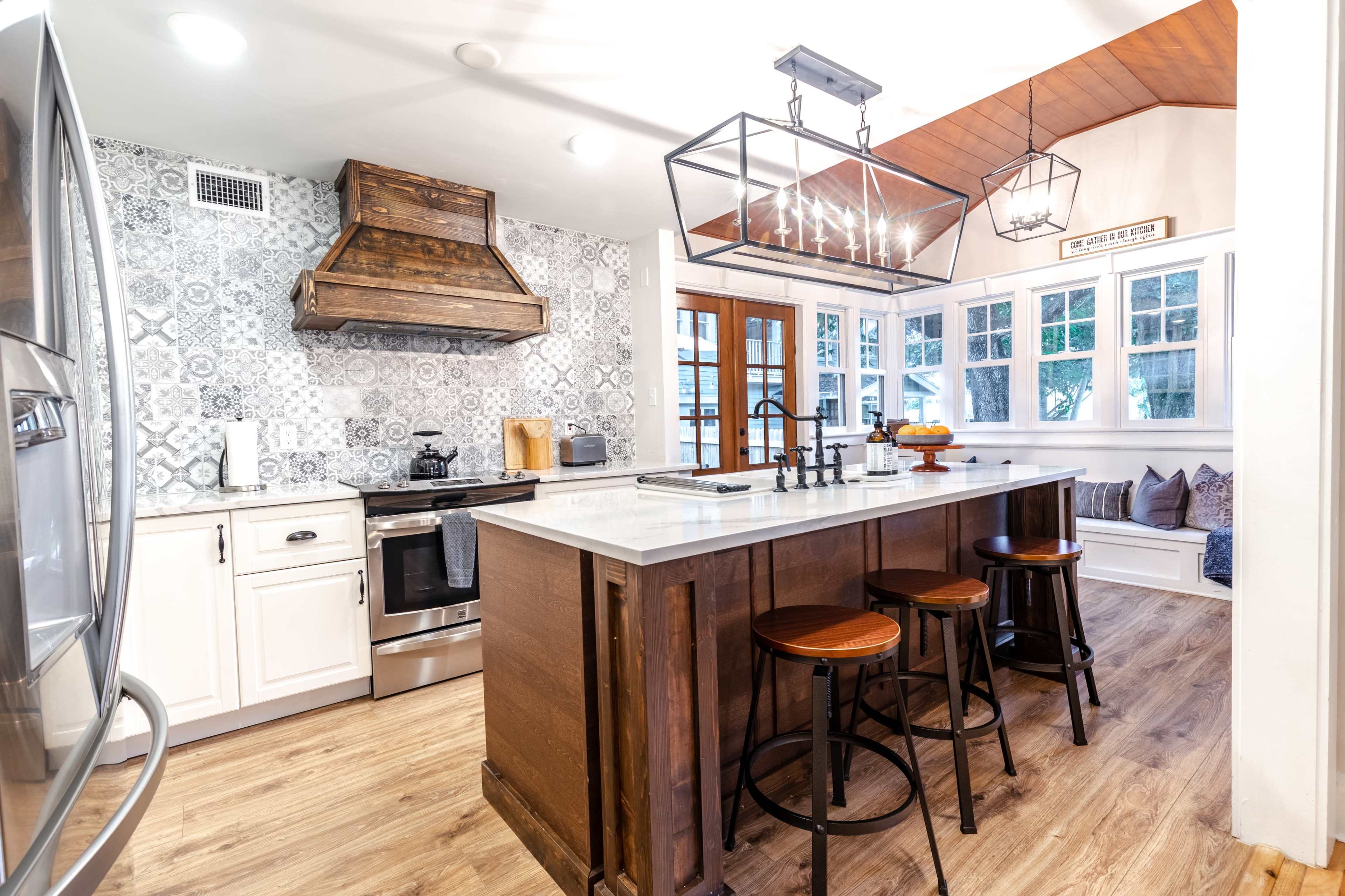A modern kitchen featuring a central island with three stools, a stainless steel stove, and patterned tile backsplash.