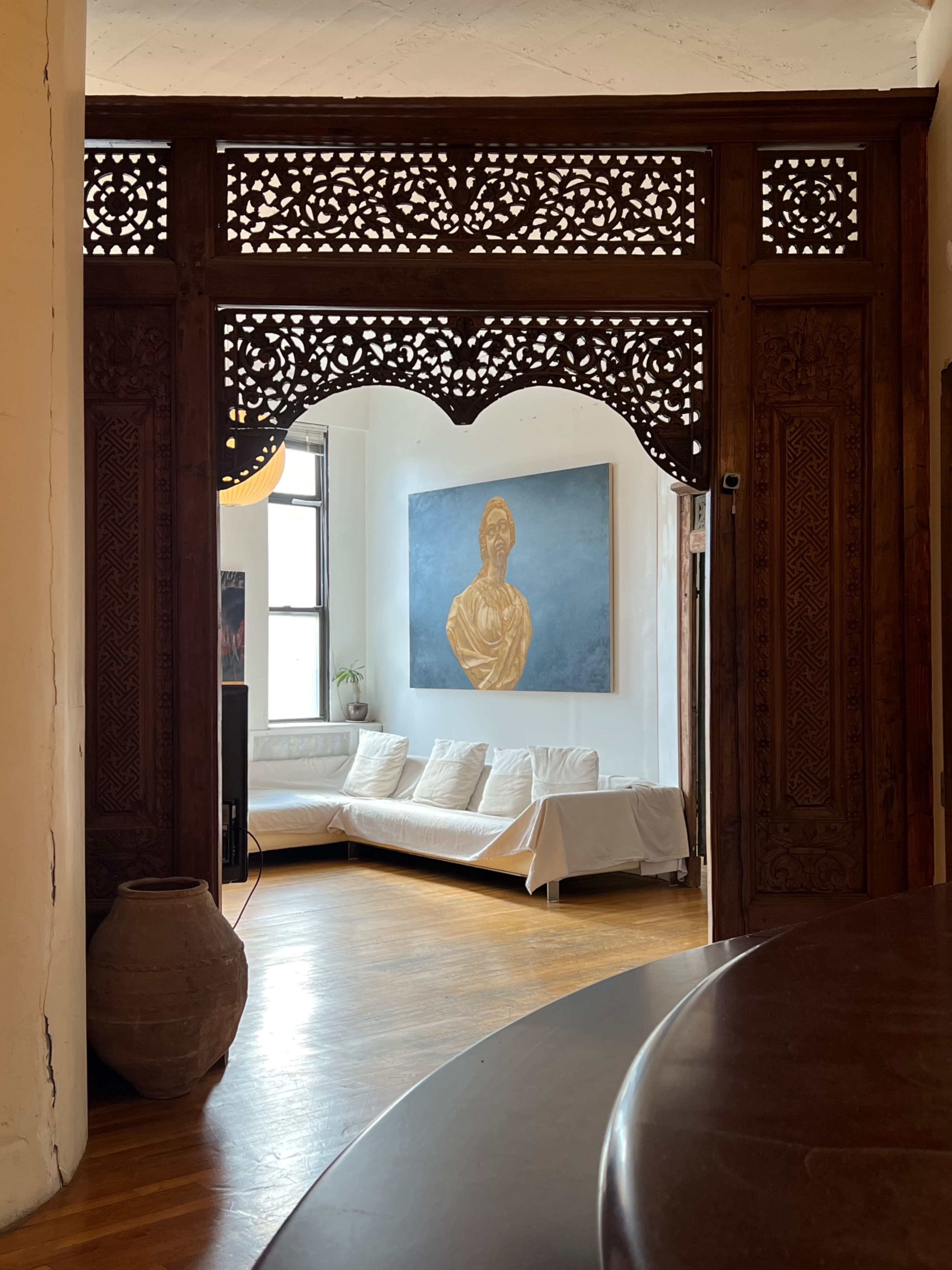 The image shows an intricately carved wooden archway leading into a bright living room with a large painting and a white sofa.