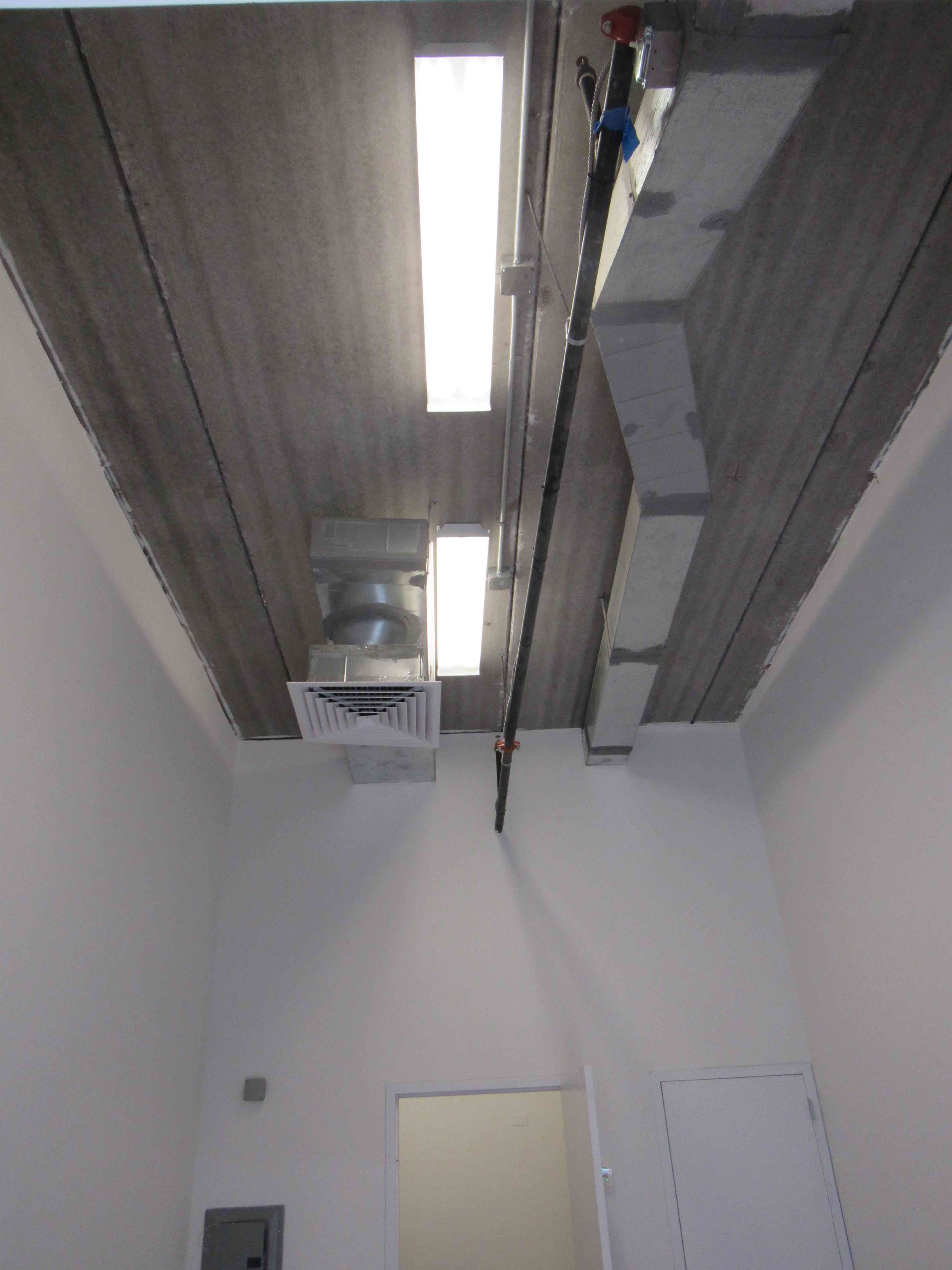 A view of a ceiling with fluorescent lights, ventilation equipment, and exposed ductwork in a plain room.