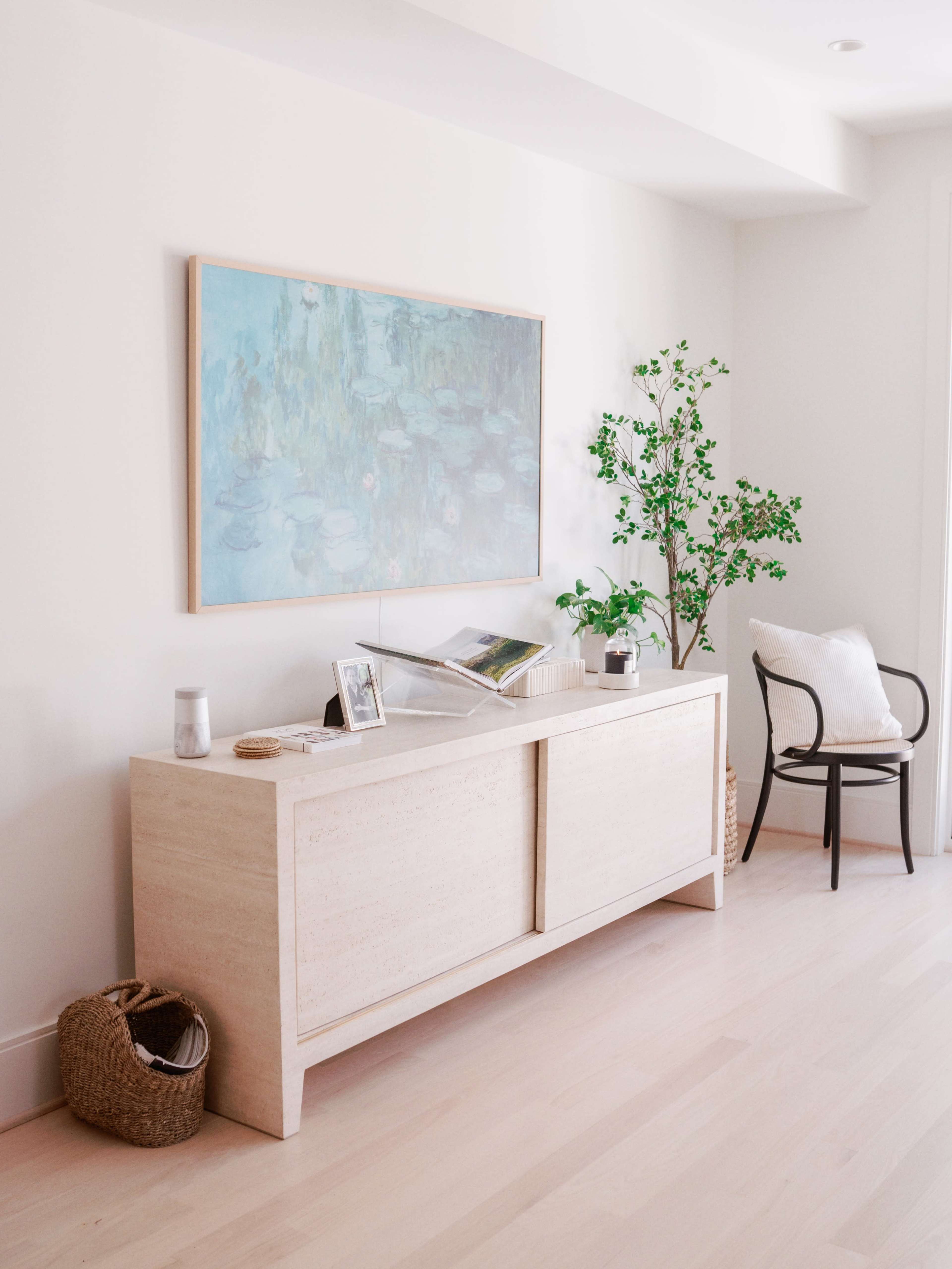 A light-colored wooden credenza against a white wall, adorned with a large painting, a potted plant, and a chair nearby.