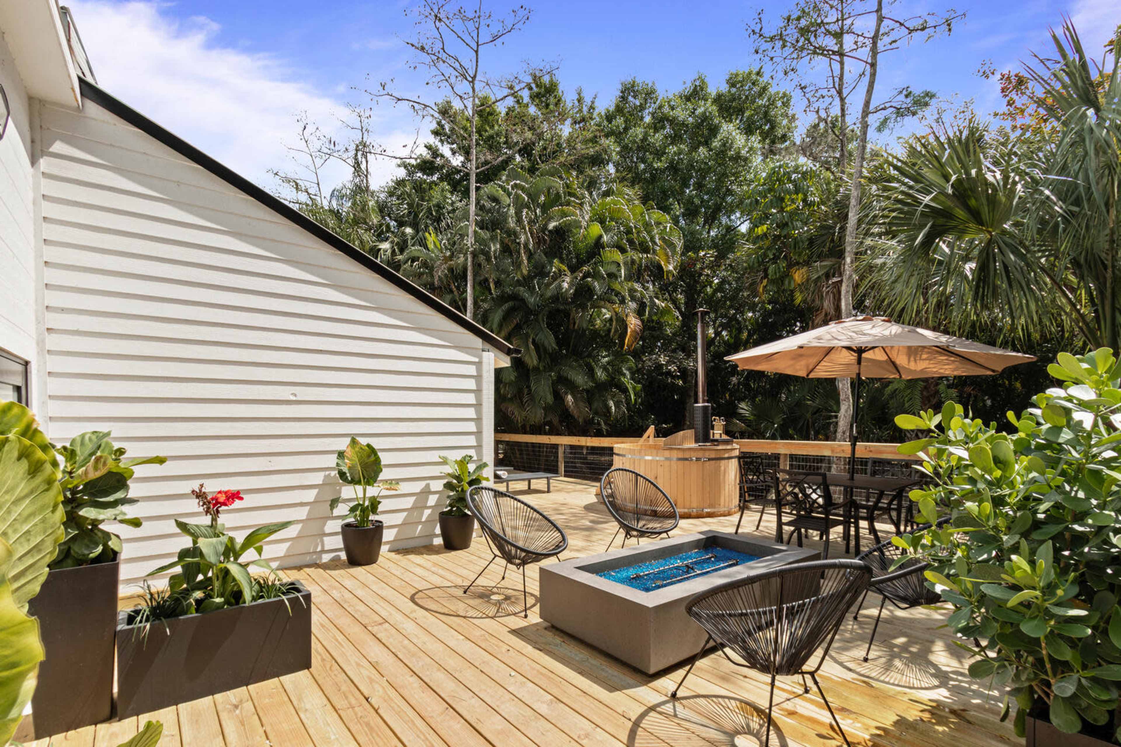 The image features a wooden deck with modern seating, a fire pit, and an umbrella-covered dining area, surrounded by lush greenery.