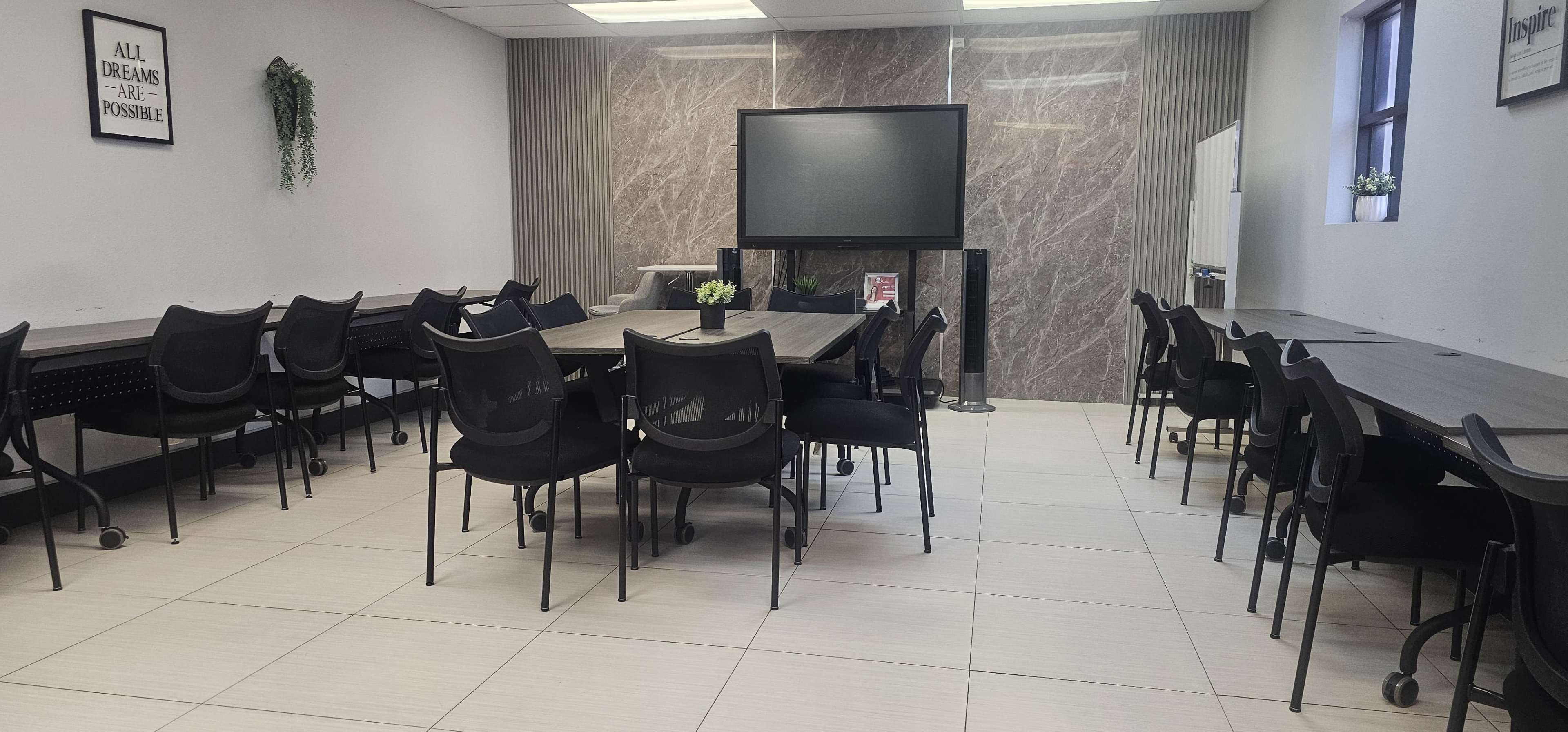 The image shows a modern room with multiple tables and chairs arranged neatly, along with a large screen at one end and decorative elements such as plants.