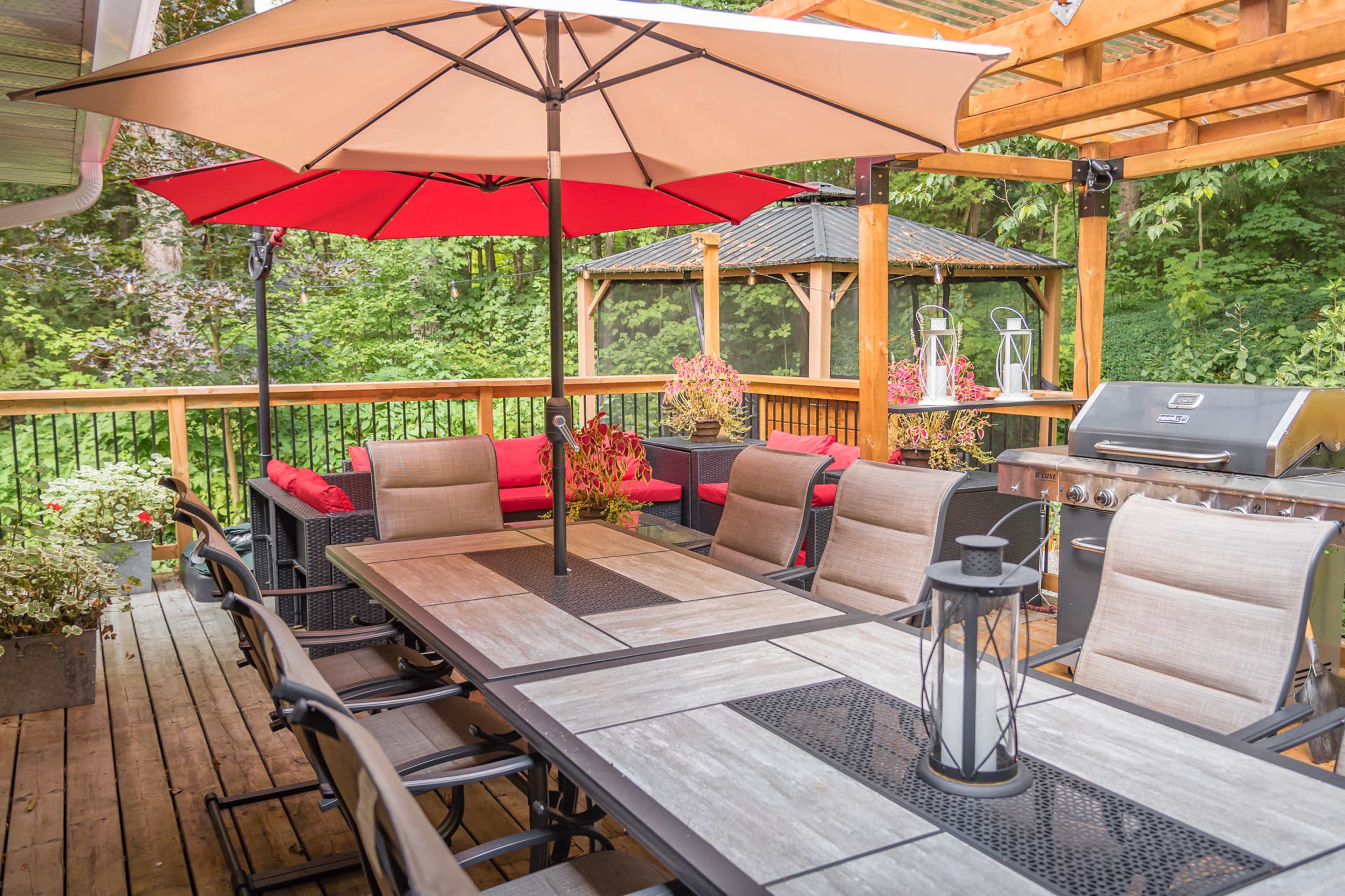 A wooden deck features a dining table with multiple chairs, an umbrella for shade, and a barbecue grill, surrounded by potted plants and a gazebo in the background.