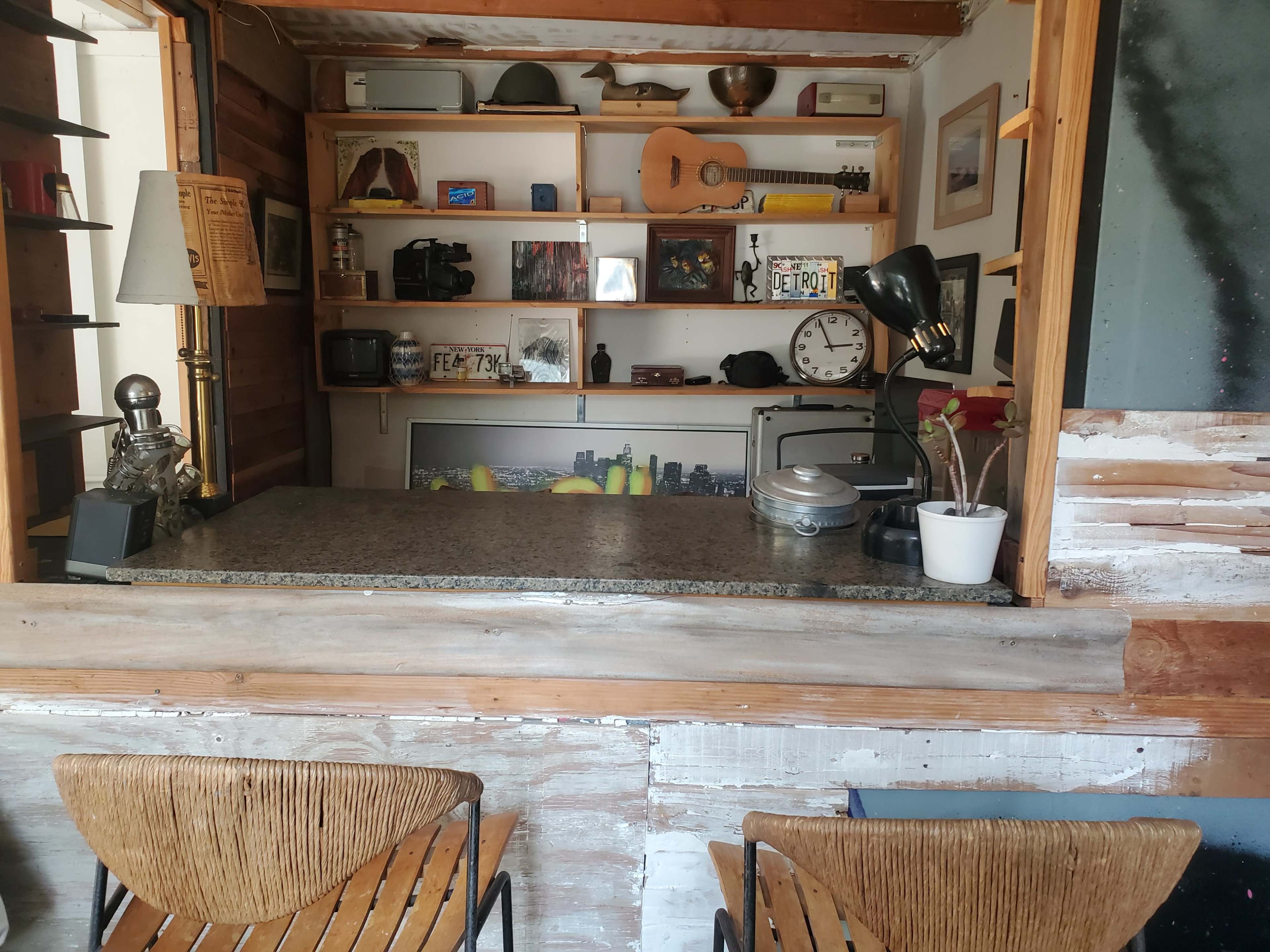 The image shows a wooden counter in a small room with shelves holding various items, including a guitar, books, and photos, and two chairs positioned in front.