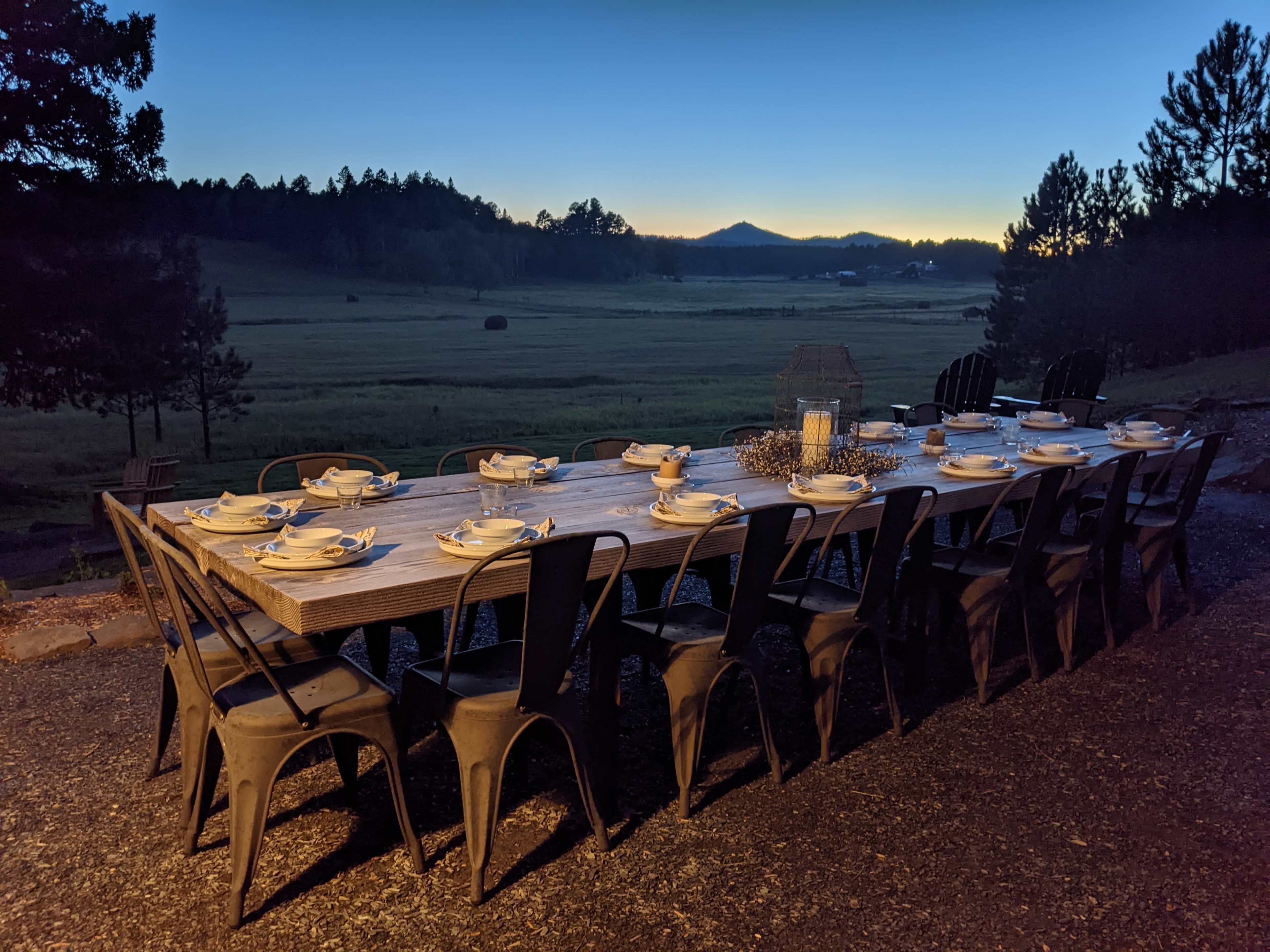 A long wooden dining table is set outdoors with plates and utensils, overlooking a grassy field at dusk.