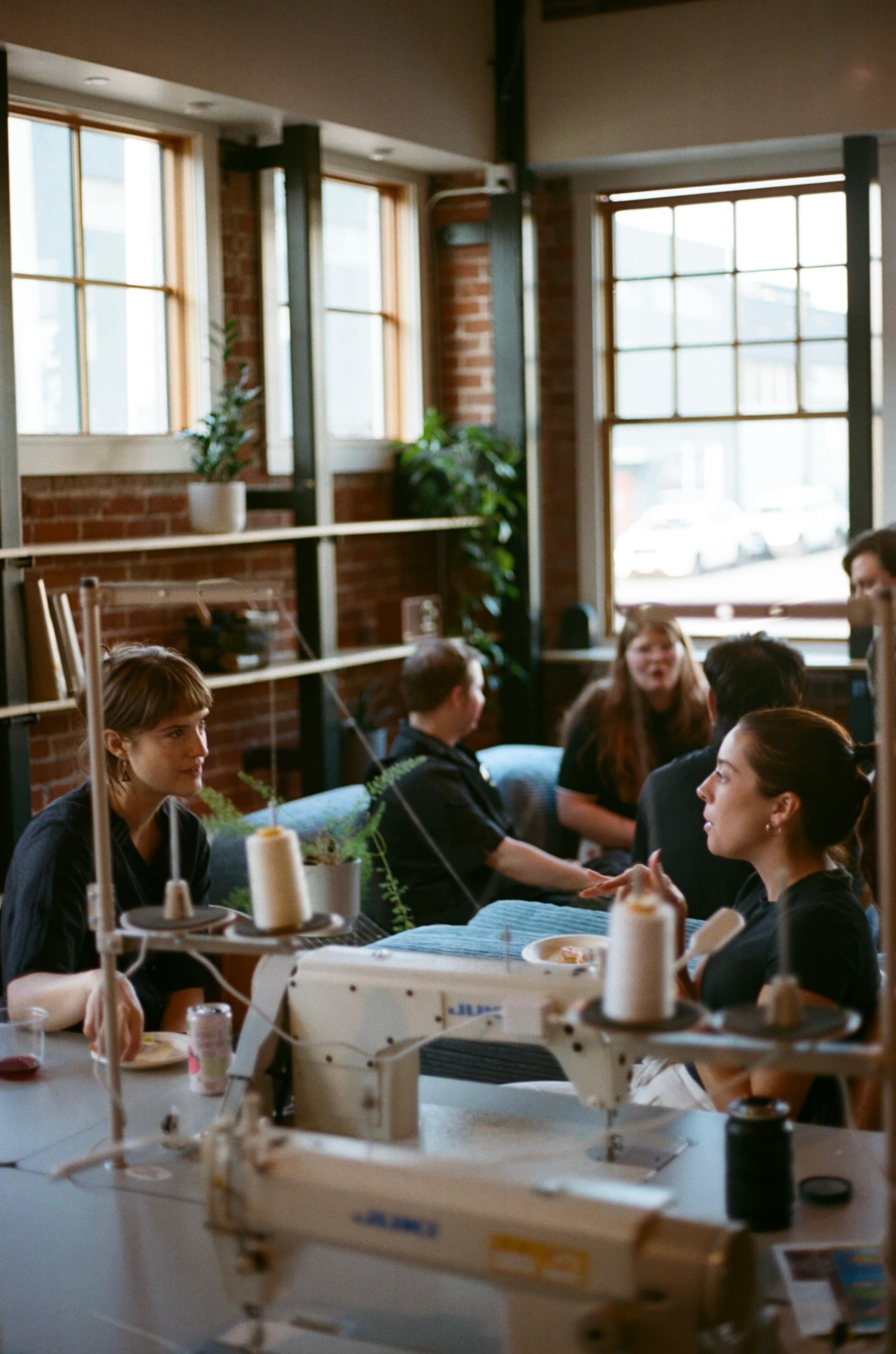 A group of people sits in a brightly lit room with sewing machines, engaging in conversation while sharing food and drinks.