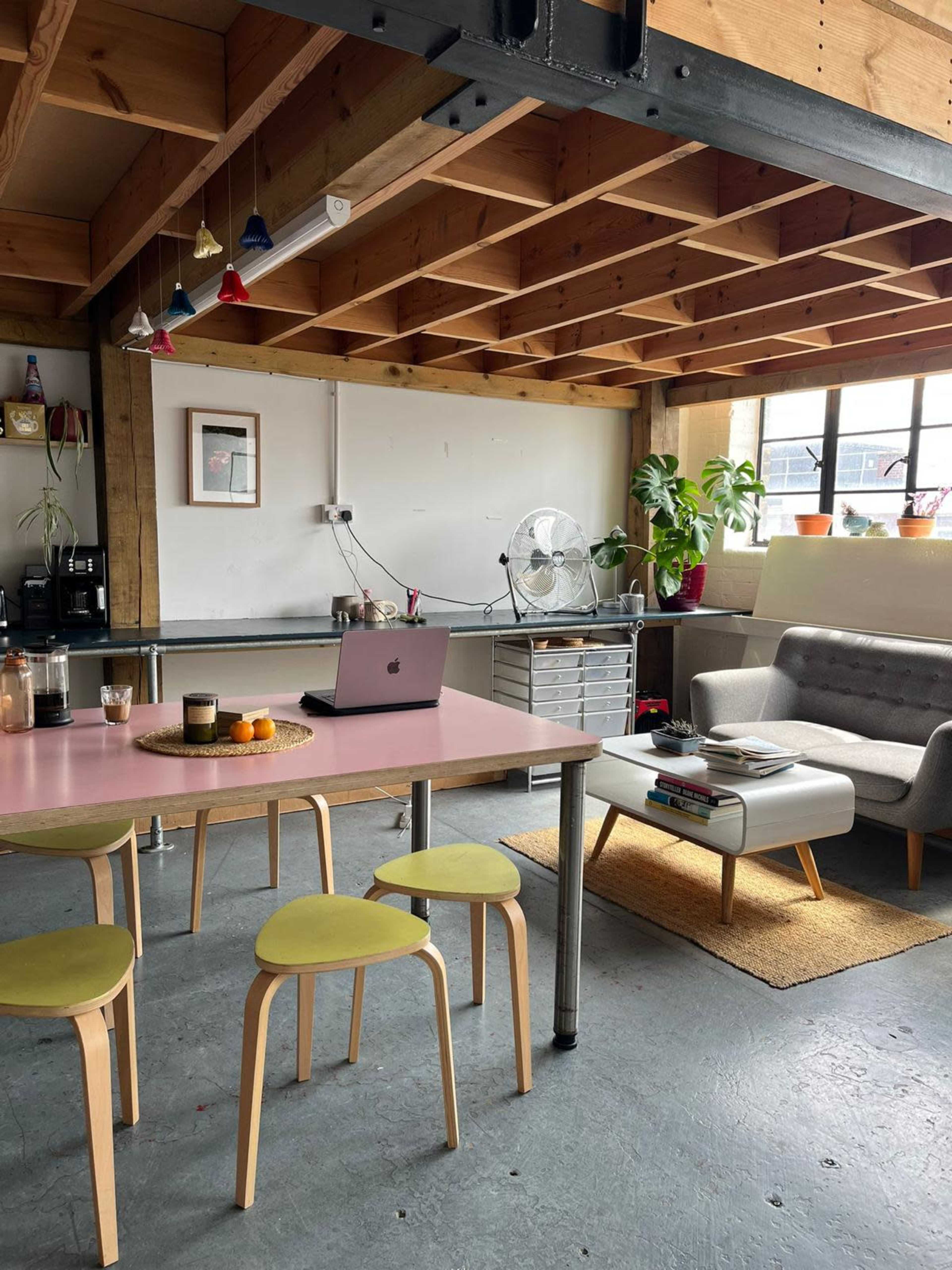 A modern interior features a dining table with yellow stools, a sofa, plants, and a fan, all under a wooden ceiling with pendant lights.