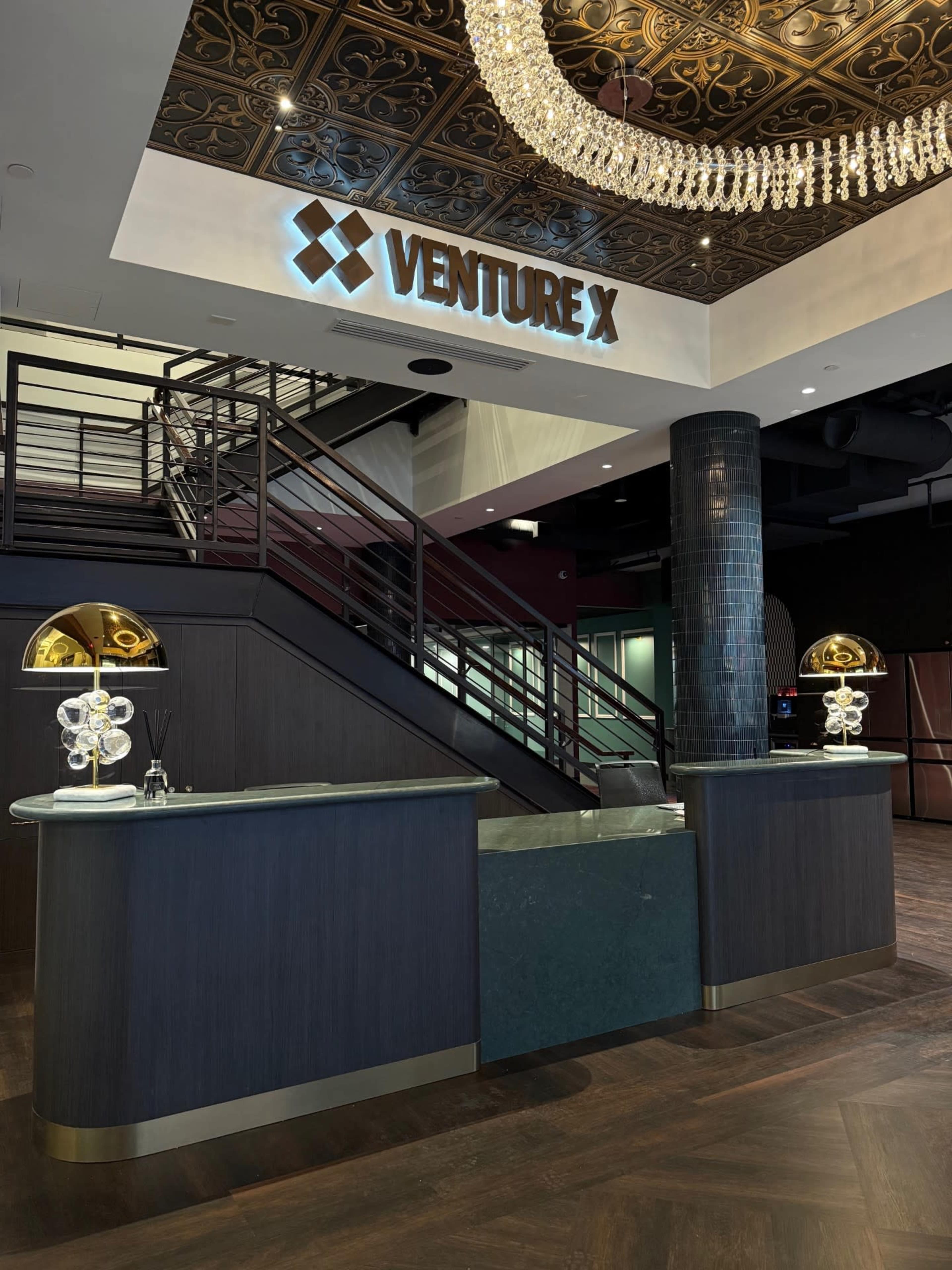 The image shows a modern reception area featuring a dark wood and marble front desk, a grand staircase, and a decorative chandelier overhead with the "VENTURE X" logo prominently displayed on the wall.