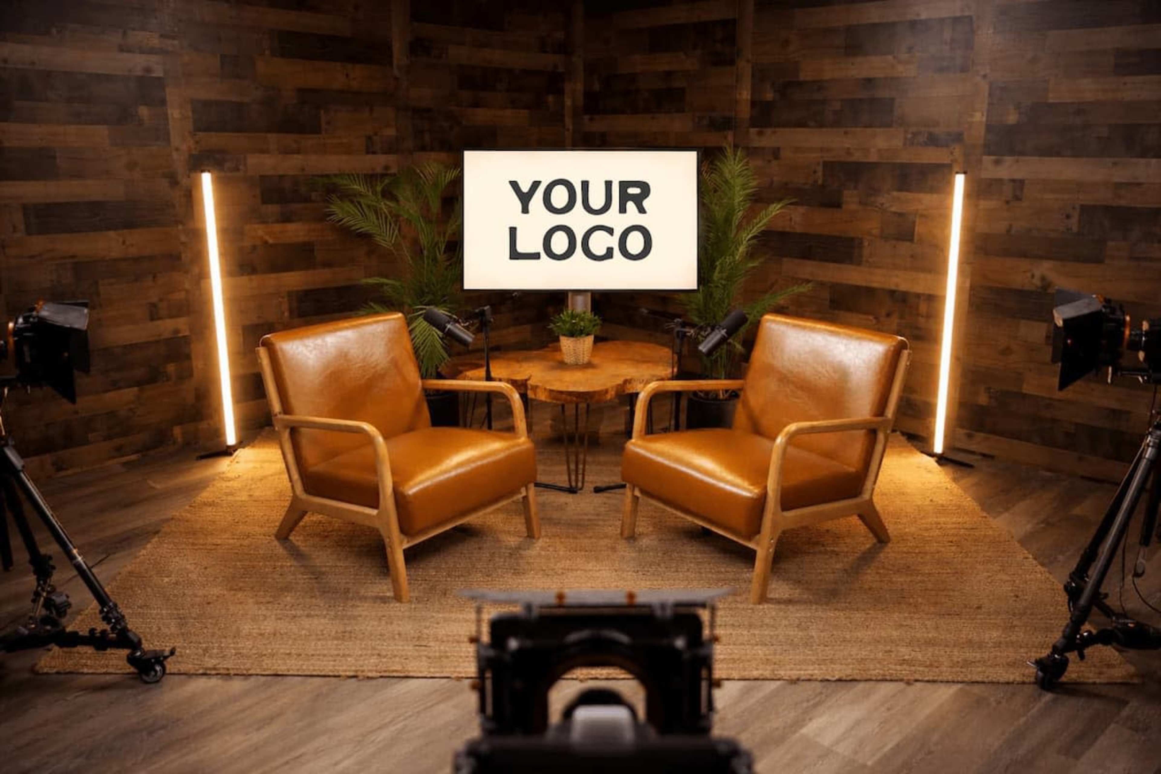 The image shows a cozy studio setup featuring two brown chairs, a wooden coffee table, and a screen displaying the text "YOUR LOGO," surrounded by plants and lighting equipment.