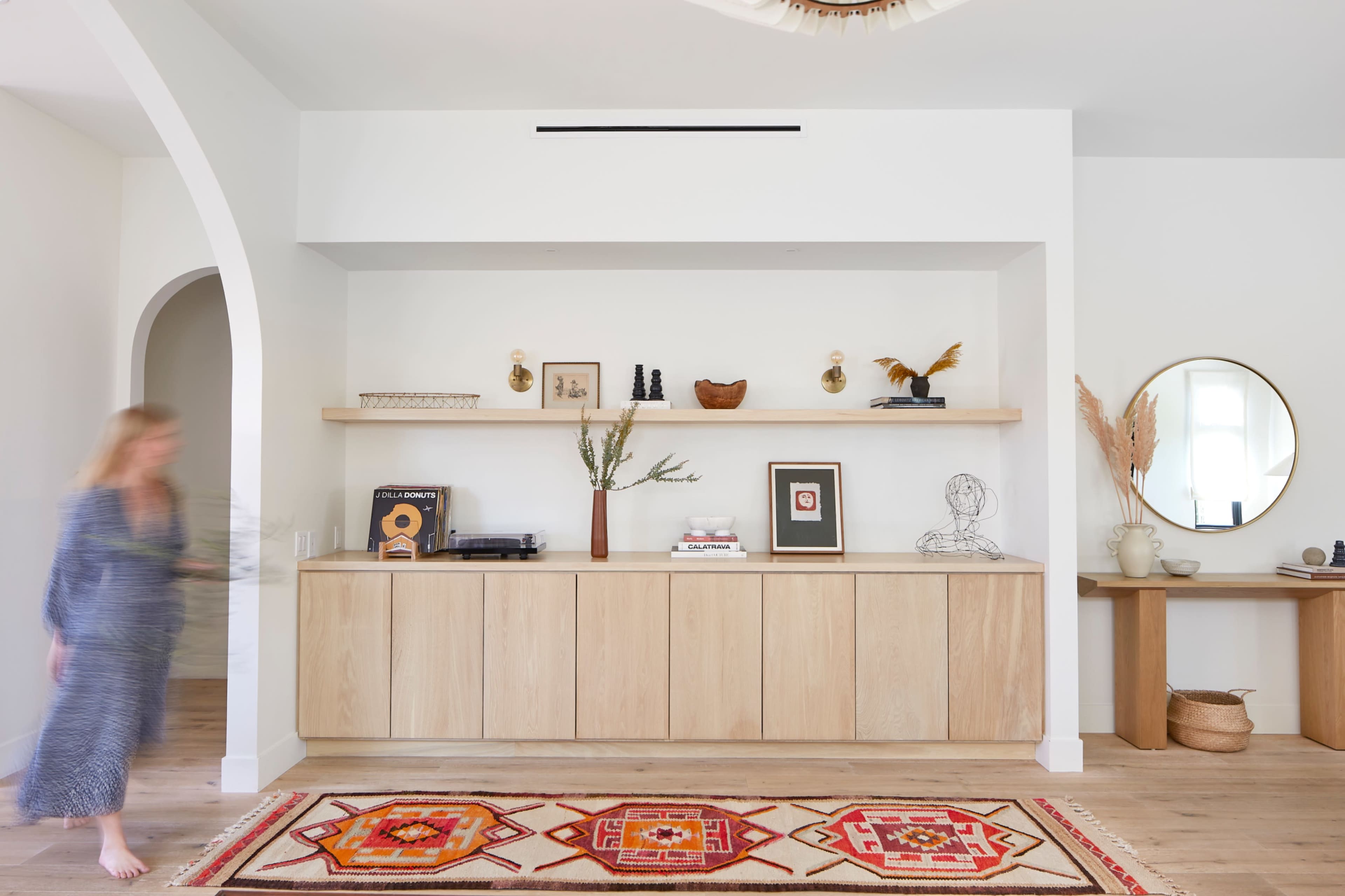 A modern living space features a wooden sideboard with decorative items, an archway, and a vibrant area rug in front of it.
