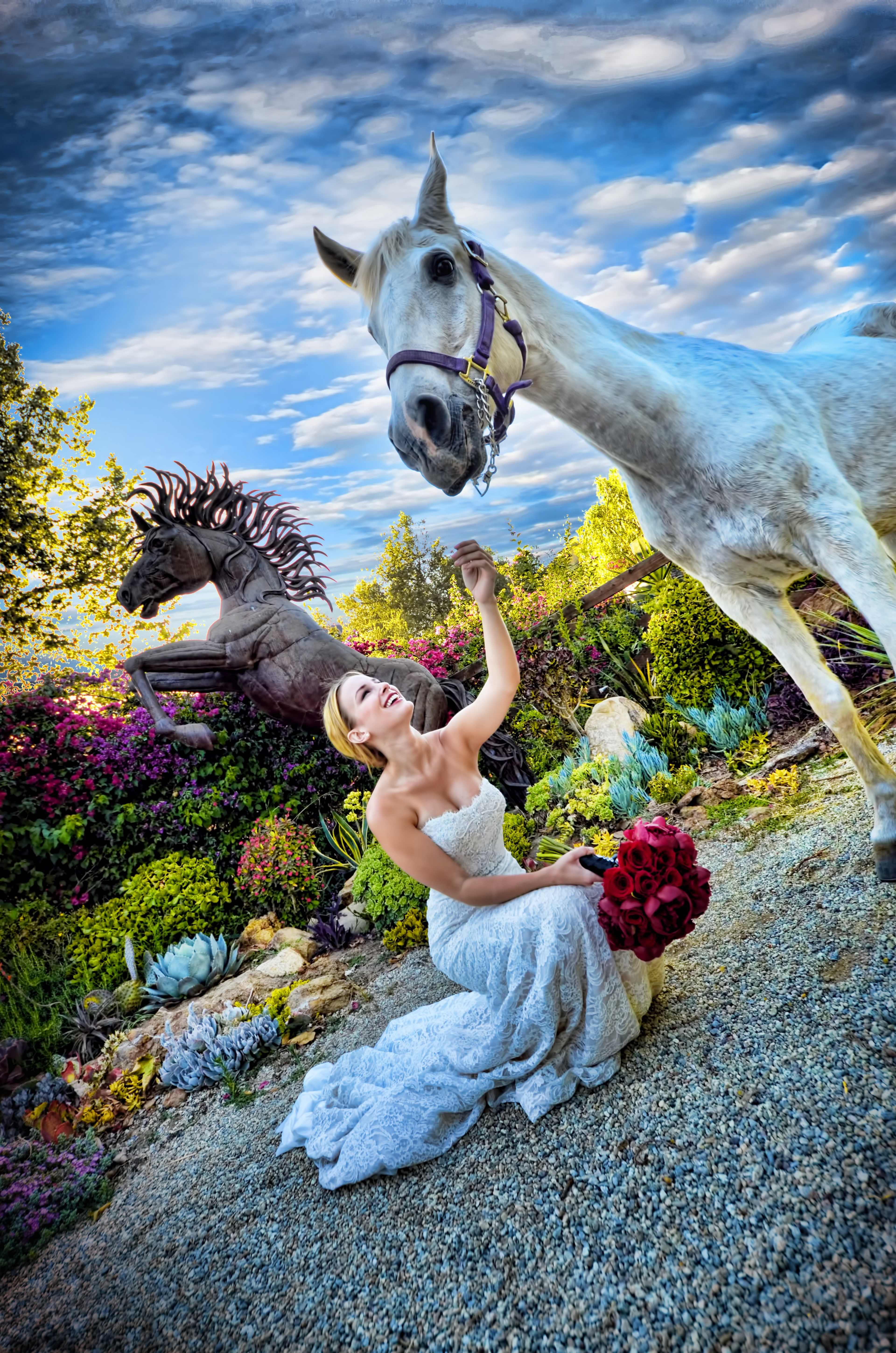 A bride in a white dress kneels in a garden while holding a bouquet of roses, reaching out to a white horse beside her, with a horse sculpture in the background.