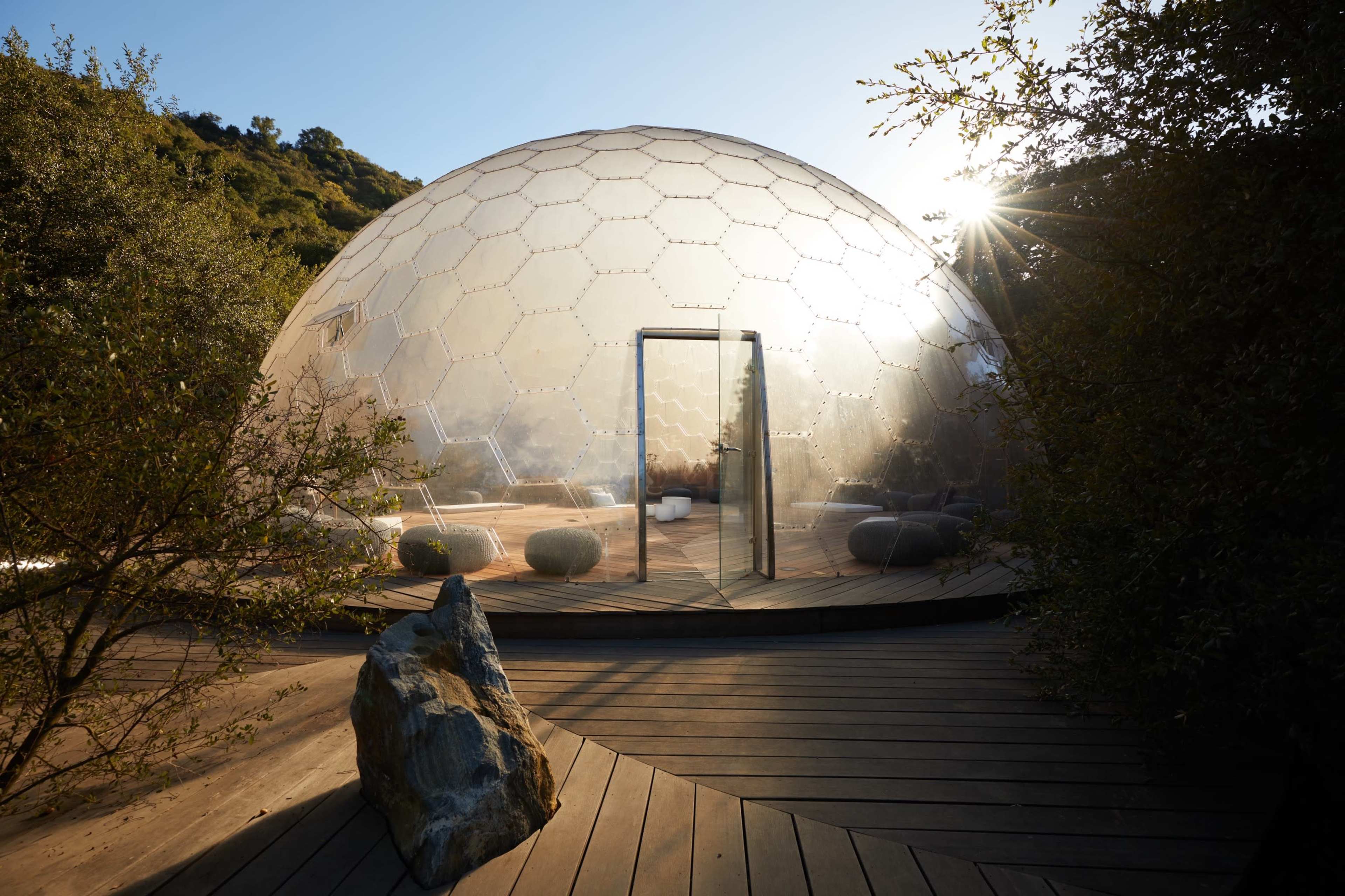 A transparent geodesic dome structure is surrounded by greenery and features a glass door opening onto a wooden deck.