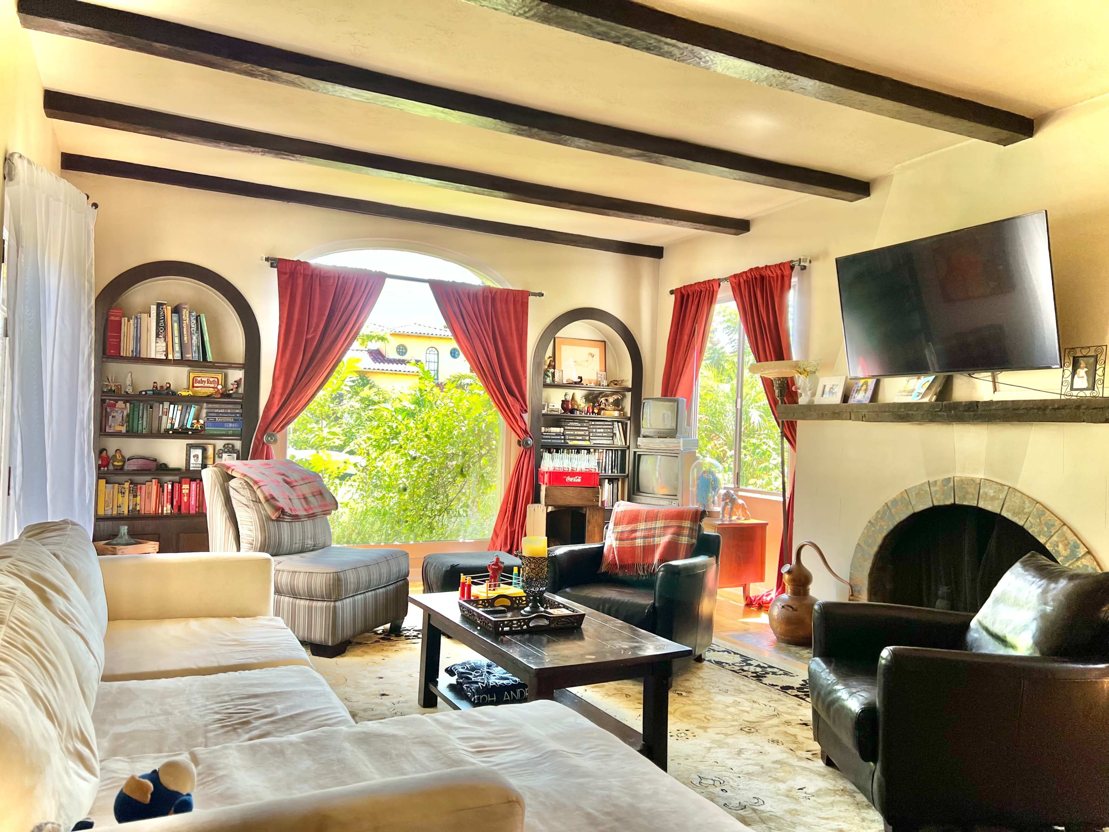 A living room features a white sofa, bookshelves filled with books, a large TV mounted on the wall, and two armchairs, all set around a central coffee table.