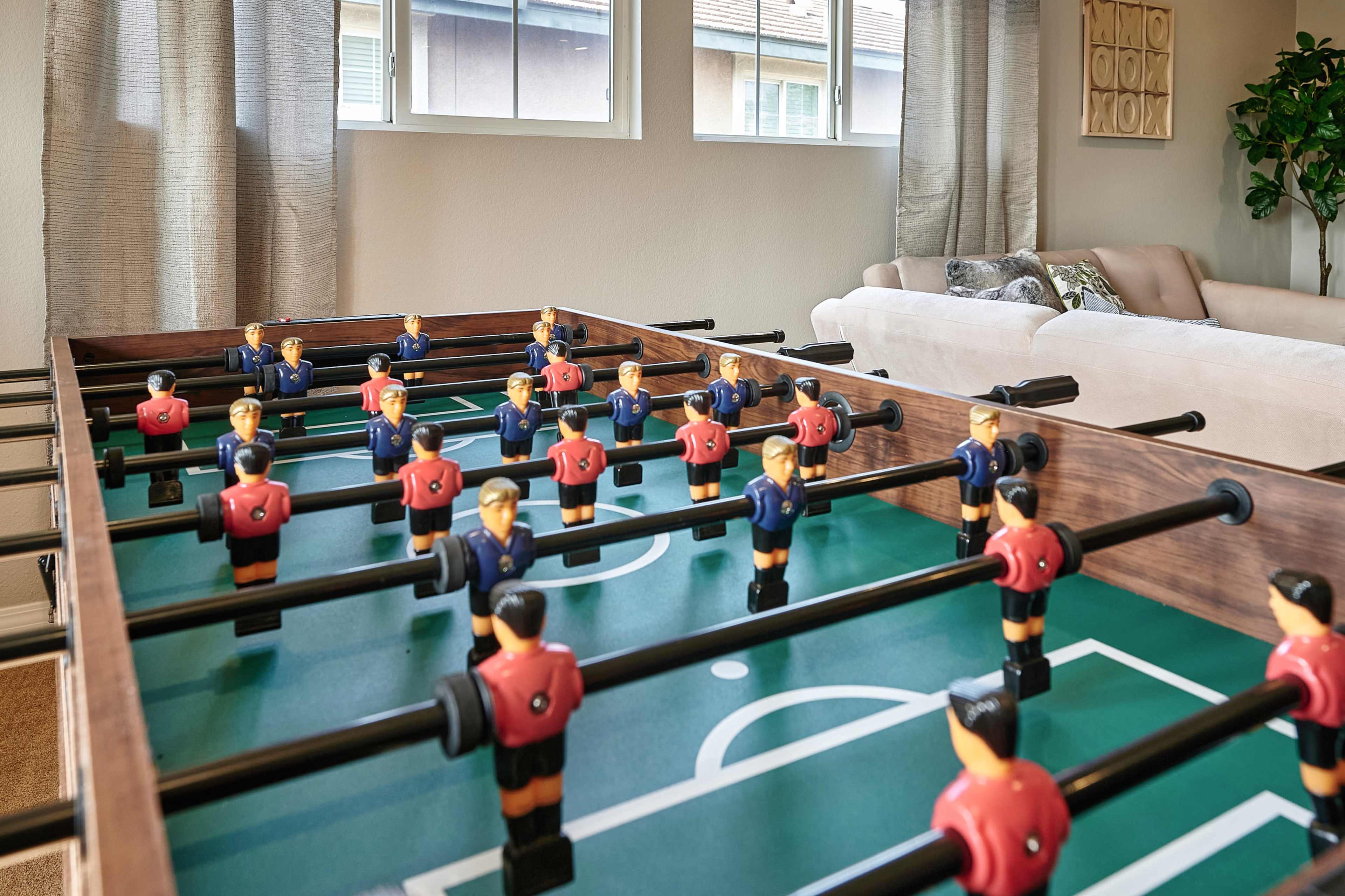A foosball table with players in red and blue jerseys is set up in a bright living room with large windows and a couch in the background.