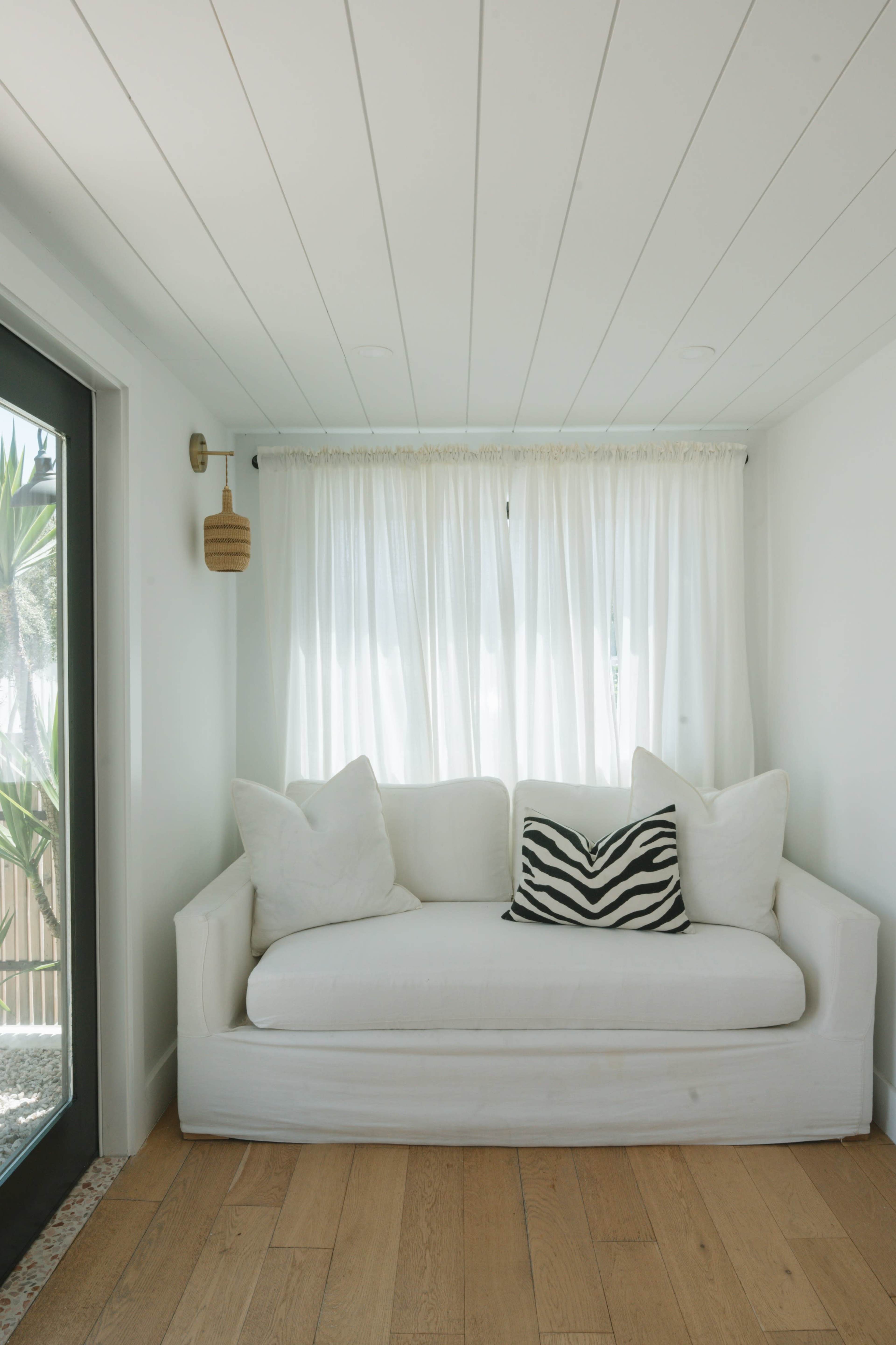 A white couch with pillows is positioned next to a window covered by sheer curtains in a bright, minimalistic room.