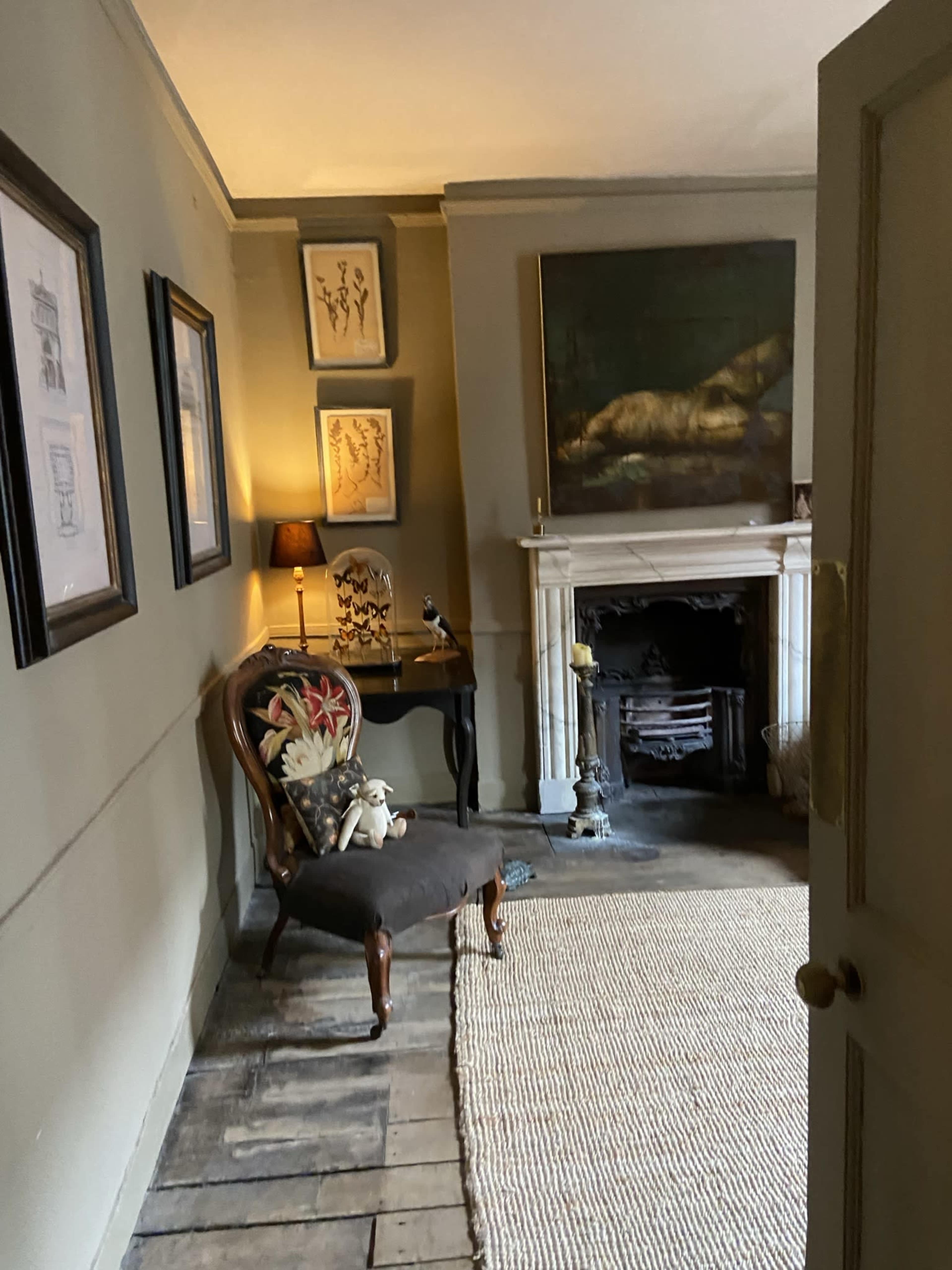 A dimly lit room with a vintage chair, a small table with a lamp, framed artwork on the walls, and a fireplace with decorative objects.