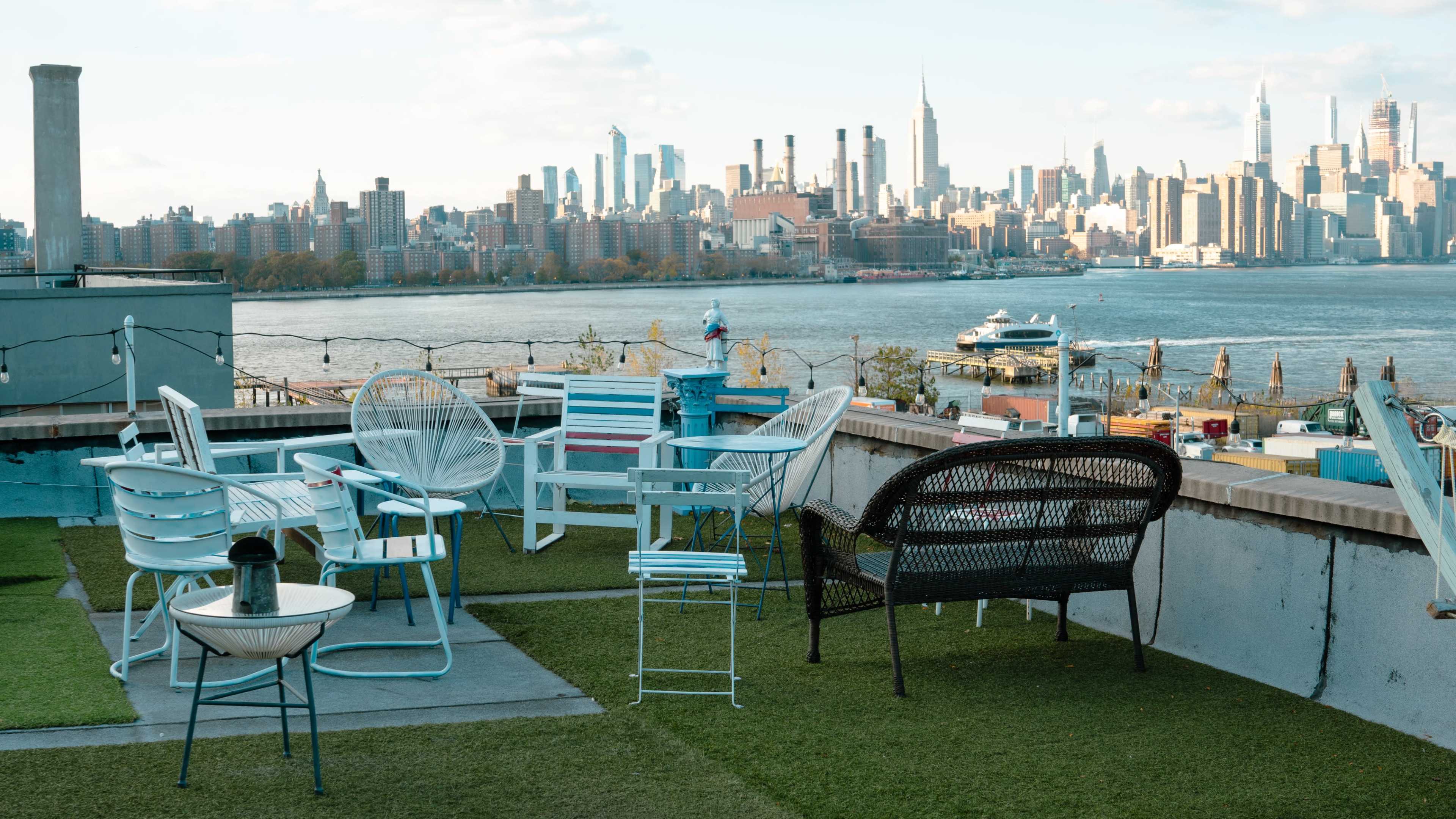The image shows a rooftop terrace with various outdoor chairs and a view of a skyline across the water.