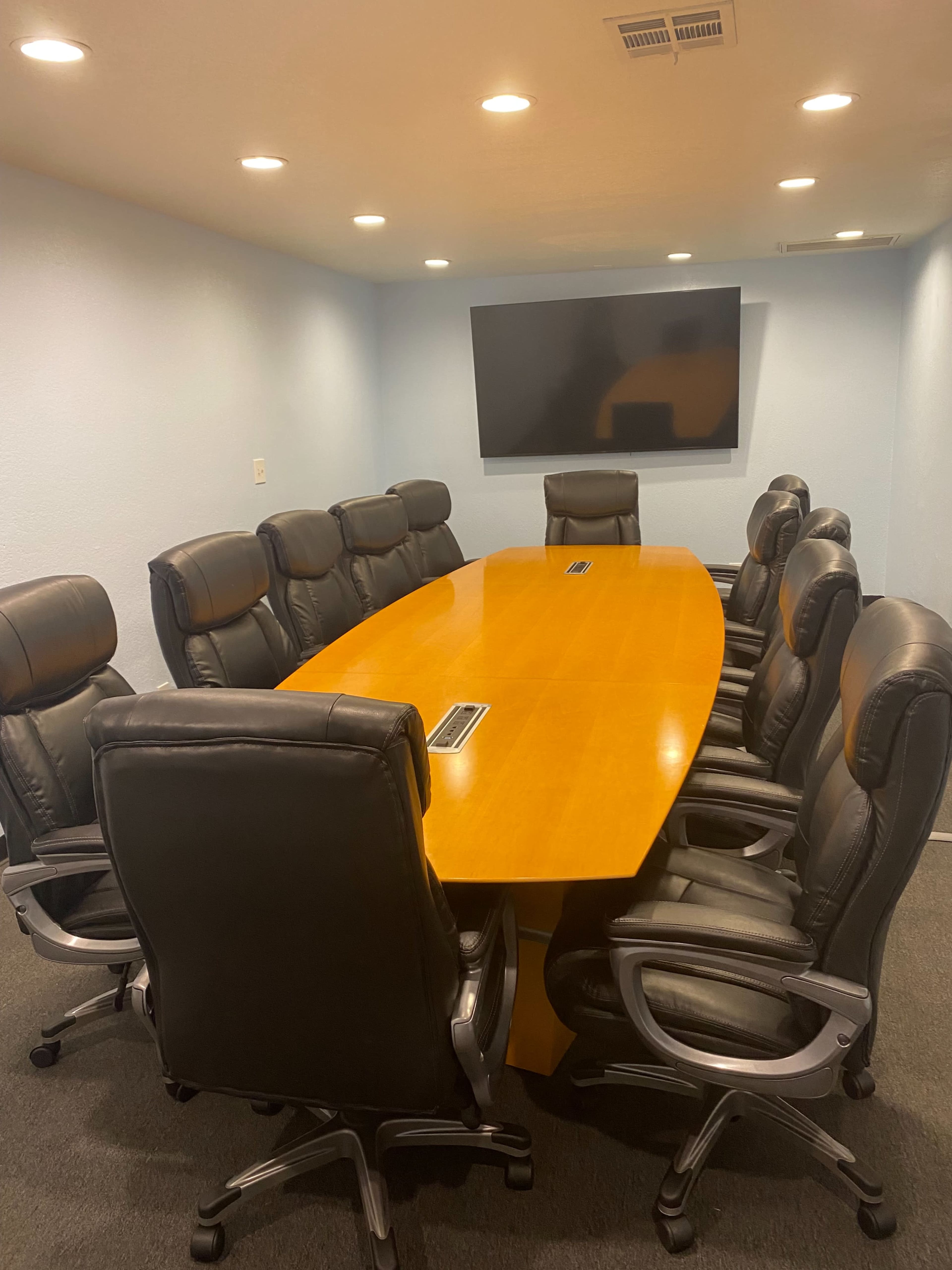 A large conference room features an oval wooden table surrounded by ten black leather chairs and a wall-mounted screen.