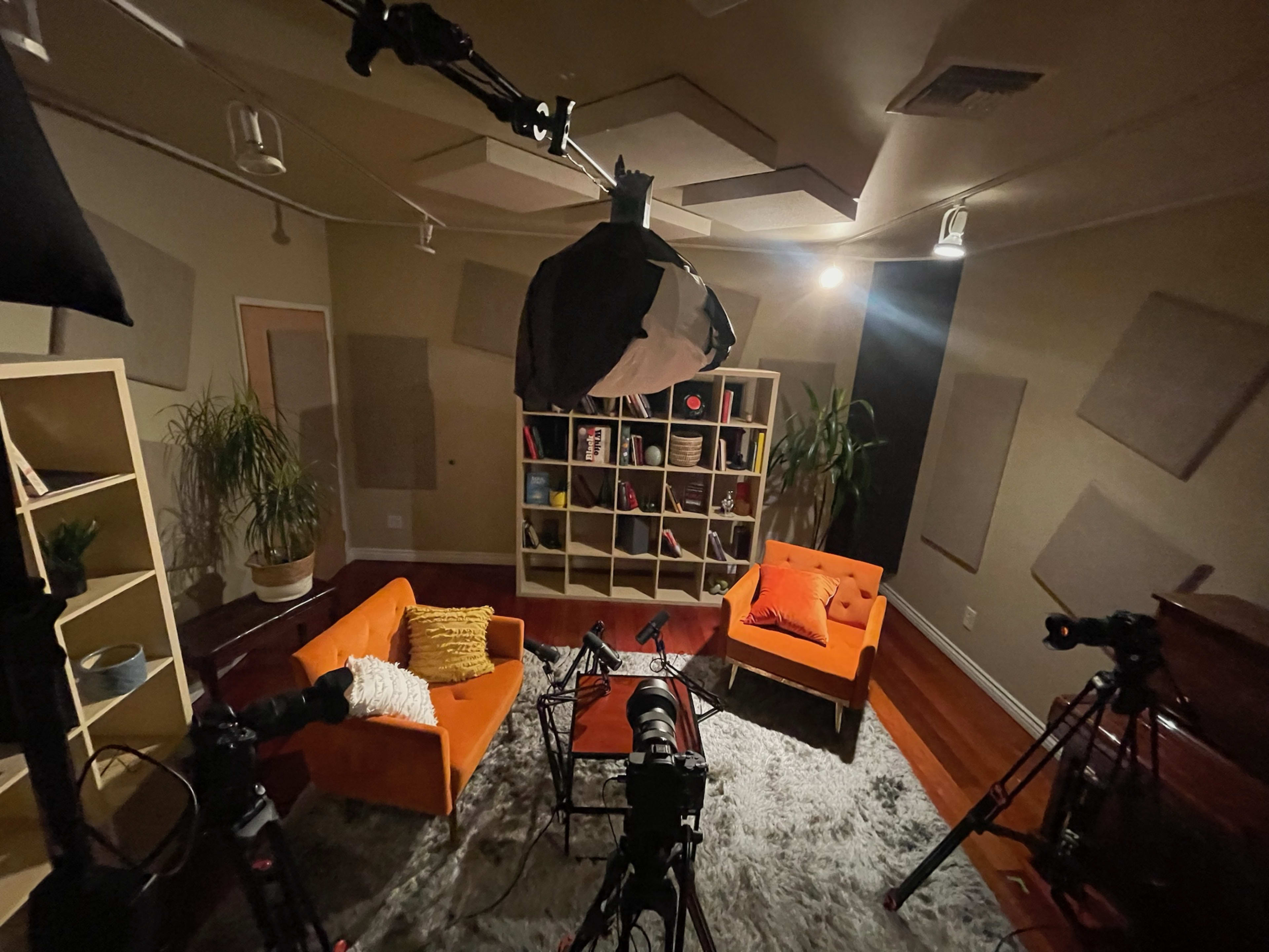 The image shows a well-lit recording studio featuring orange furniture, a shelving unit filled with books and decorative items, and multiple cameras set up for video production.