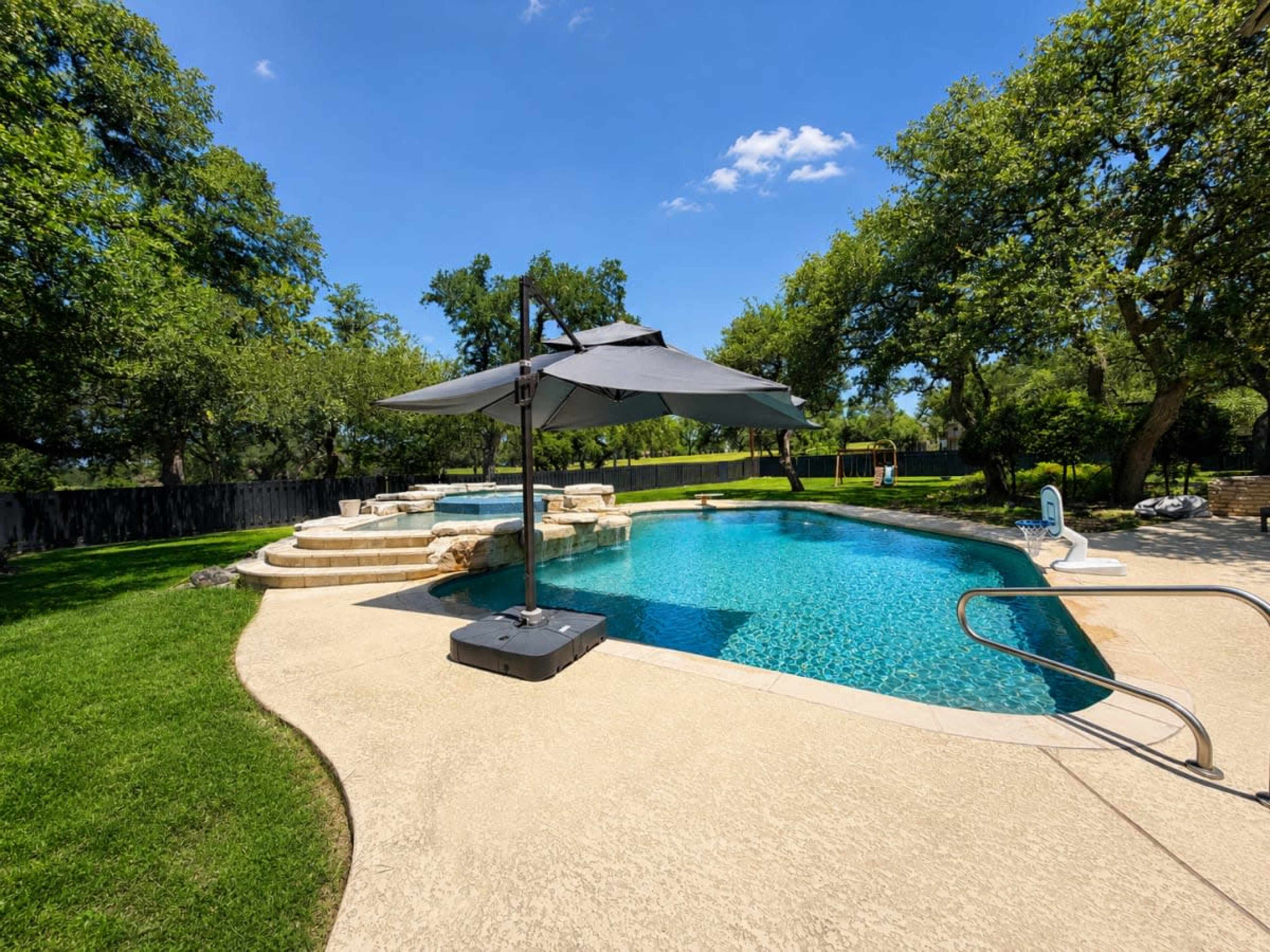 The image shows a backyard swimming pool with a surrounding deck, a shaded area from a large umbrella, and landscaped greenery in the background.