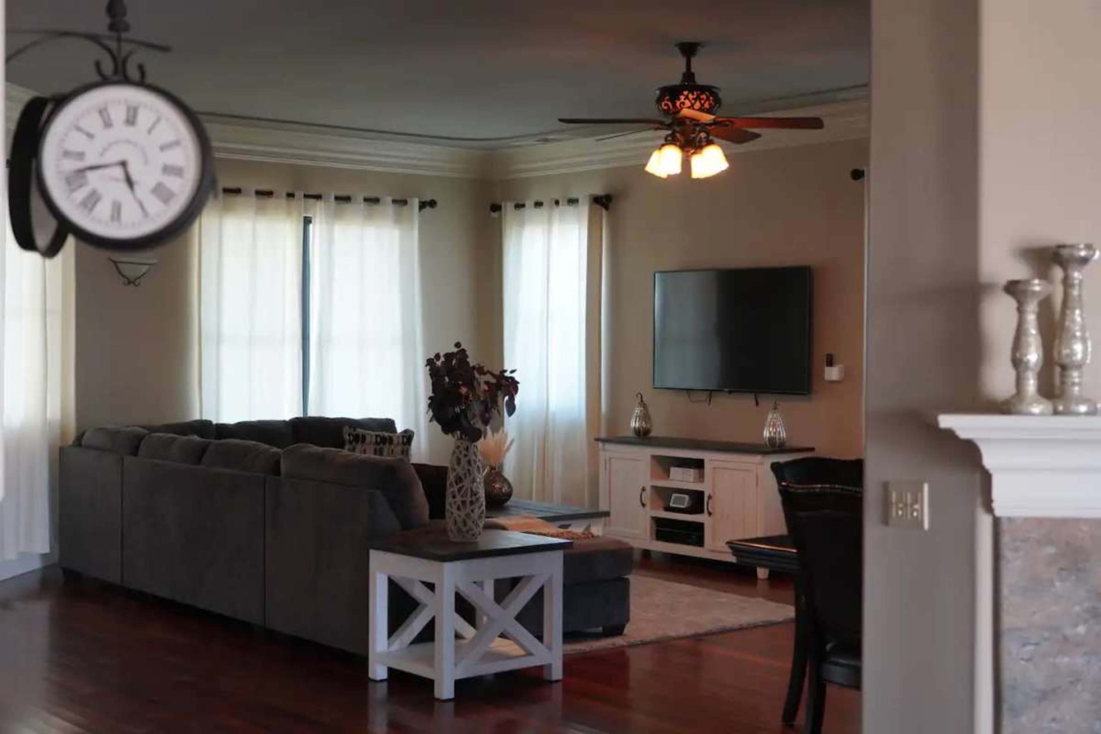 The image features a living room with a gray sectional sofa, a white television stand, and a wall-mounted TV, illuminated by natural light filtering through sheer curtains.