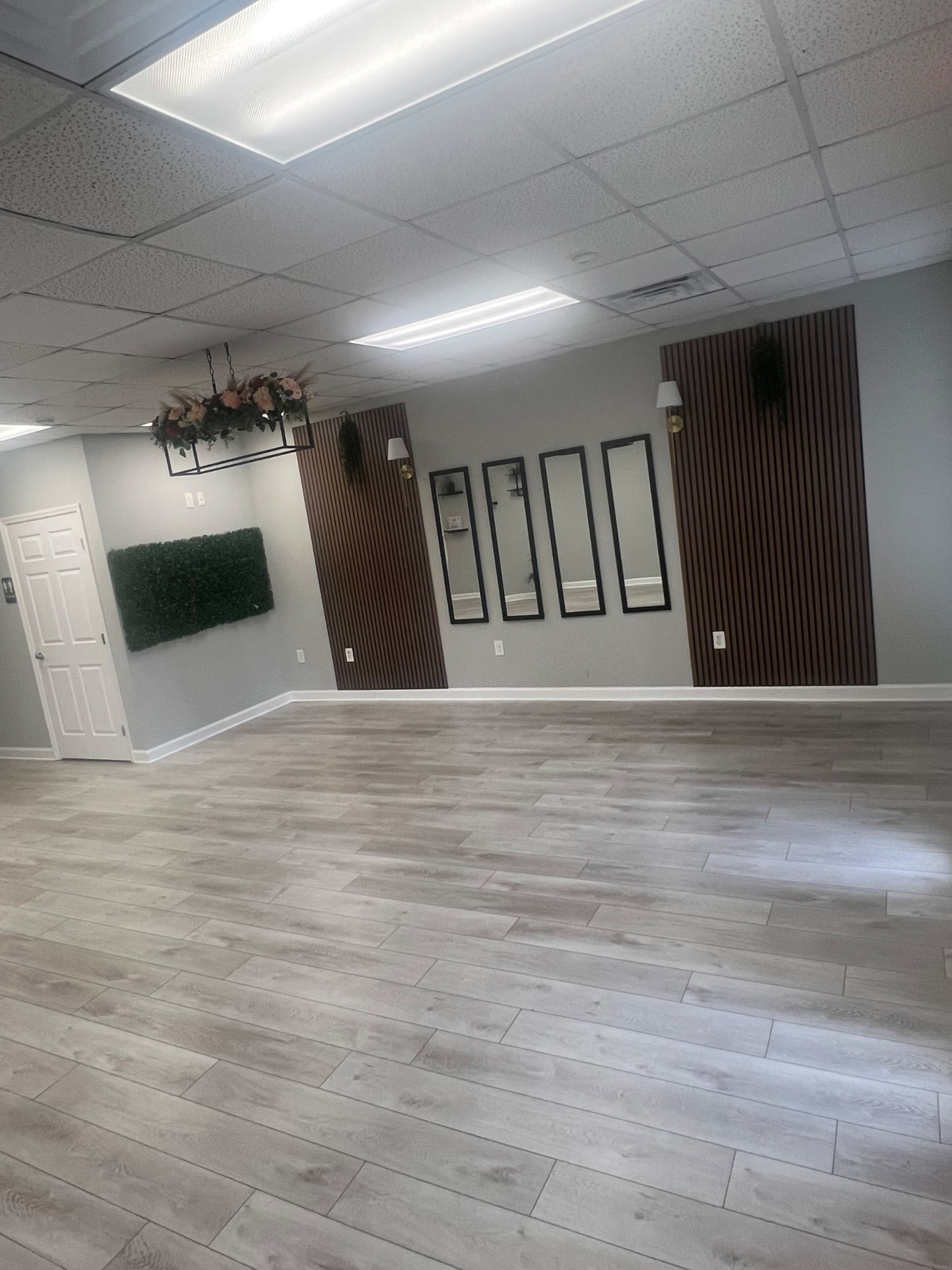 The image depicts a spacious, blank interior room with wooden flooring, light gray walls, and decorative elements including a plant wall and three vertical mirrors.
