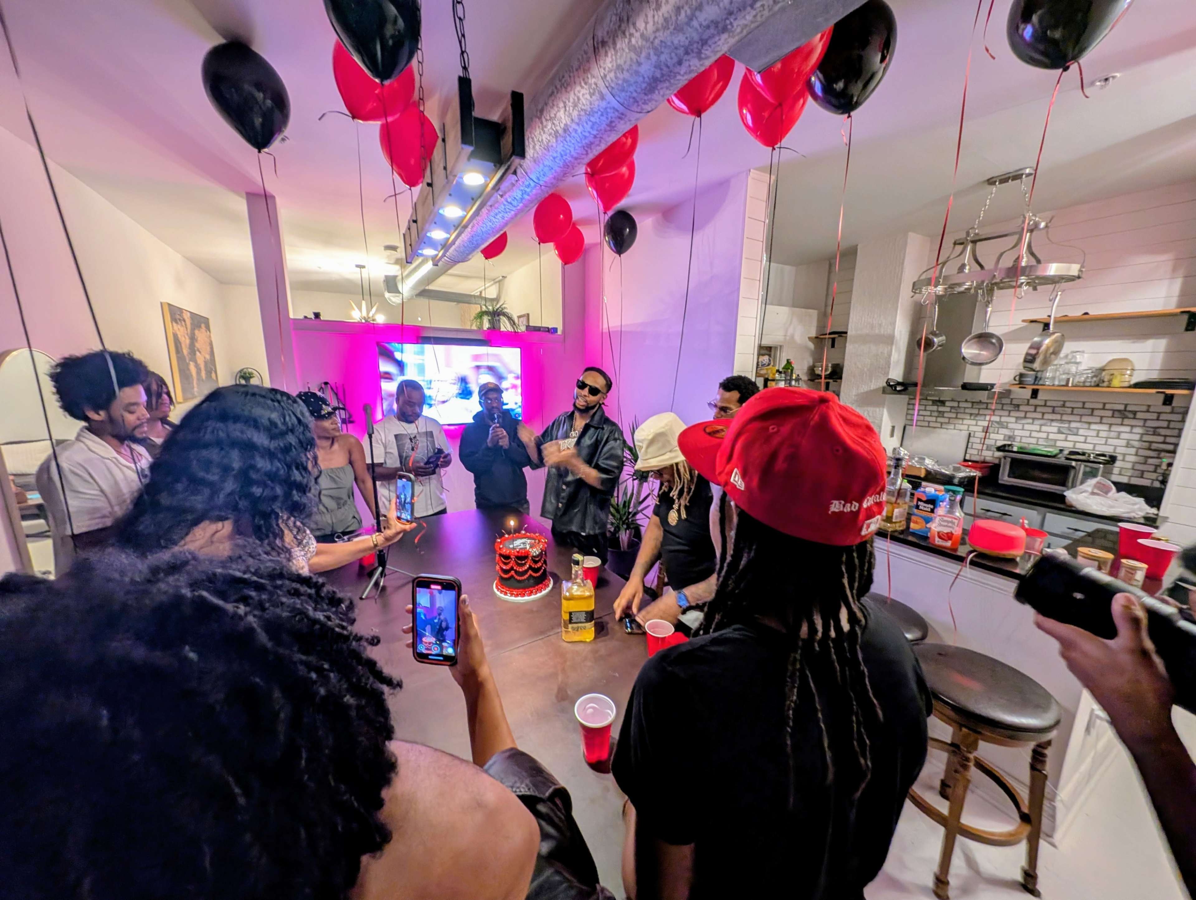 A group of people gathers in a kitchen decorated with red and black balloons, celebrating with drinks, a cake, and a screen displaying visuals in the background.