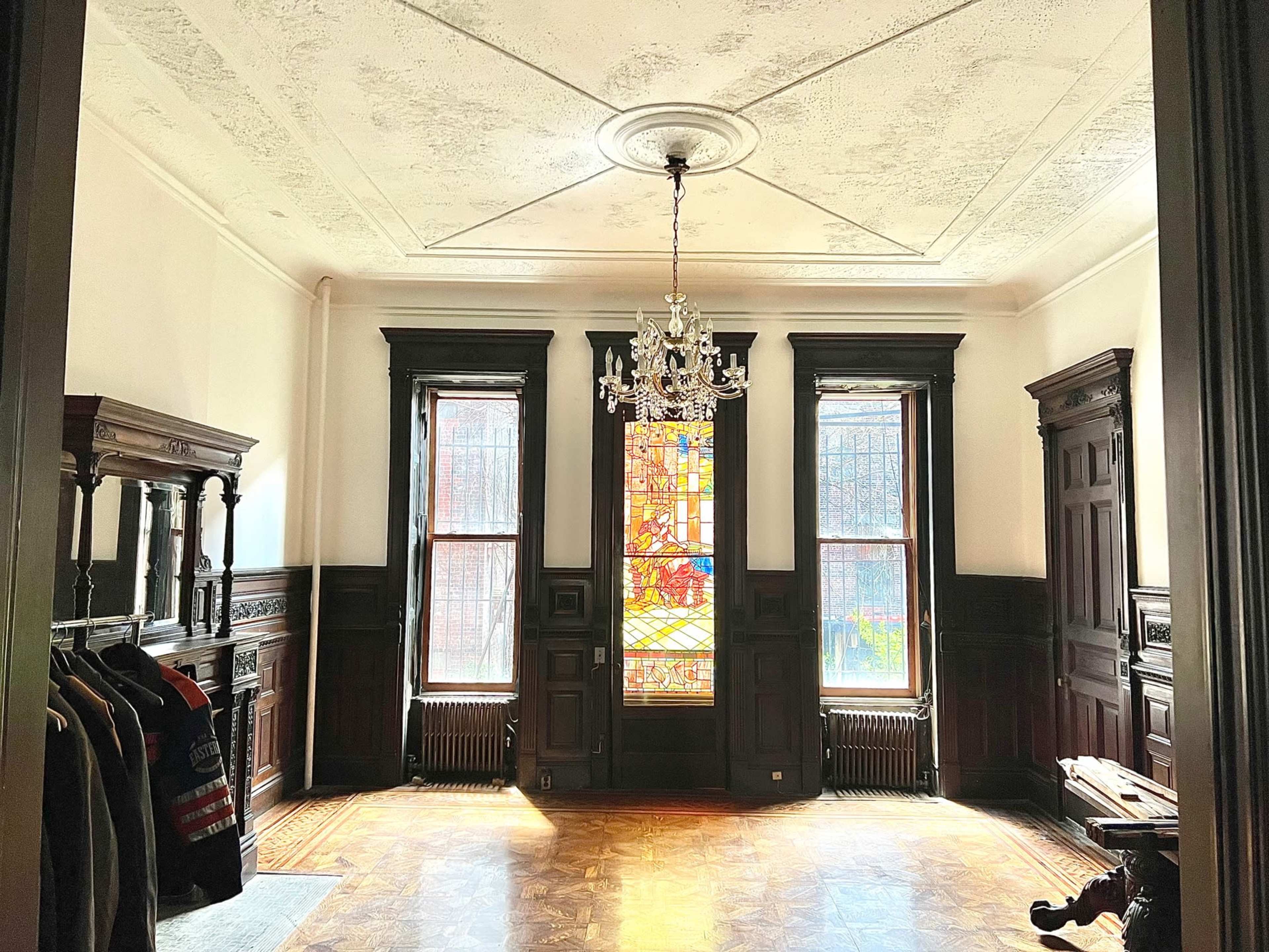 The image shows a spacious, empty room featuring dark wooden trim, large windows with stained glass, a chandelier, and wooden furniture along the walls.
