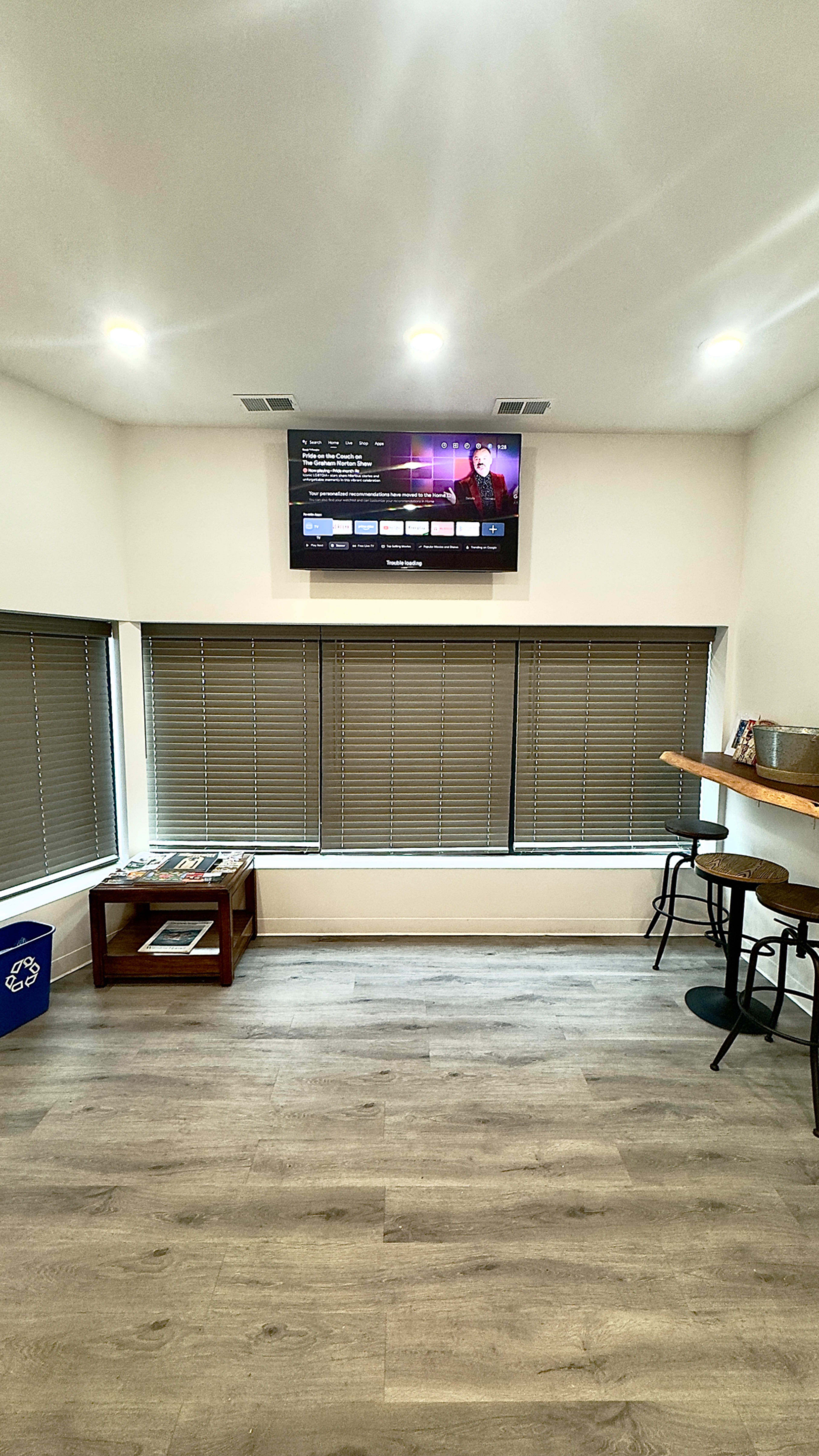 The image shows a room with wooden flooring, two large windows covered by horizontal blinds, a wall-mounted television displaying content, and a small bar area with two stools.
