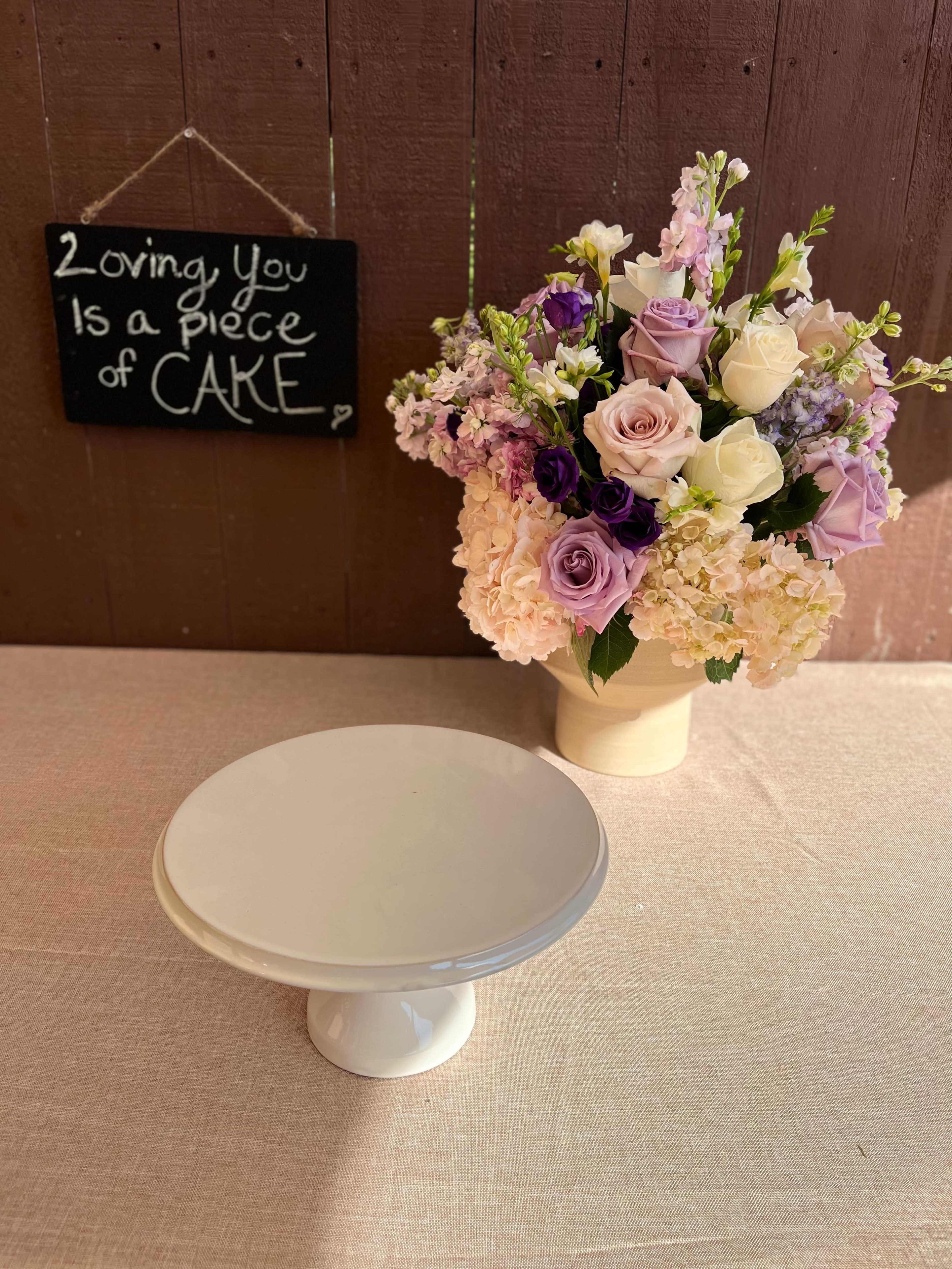 A white cake stand is positioned next to a floral arrangement, while a chalkboard sign above reads, "Loving You Is a Piece of CAKE."
